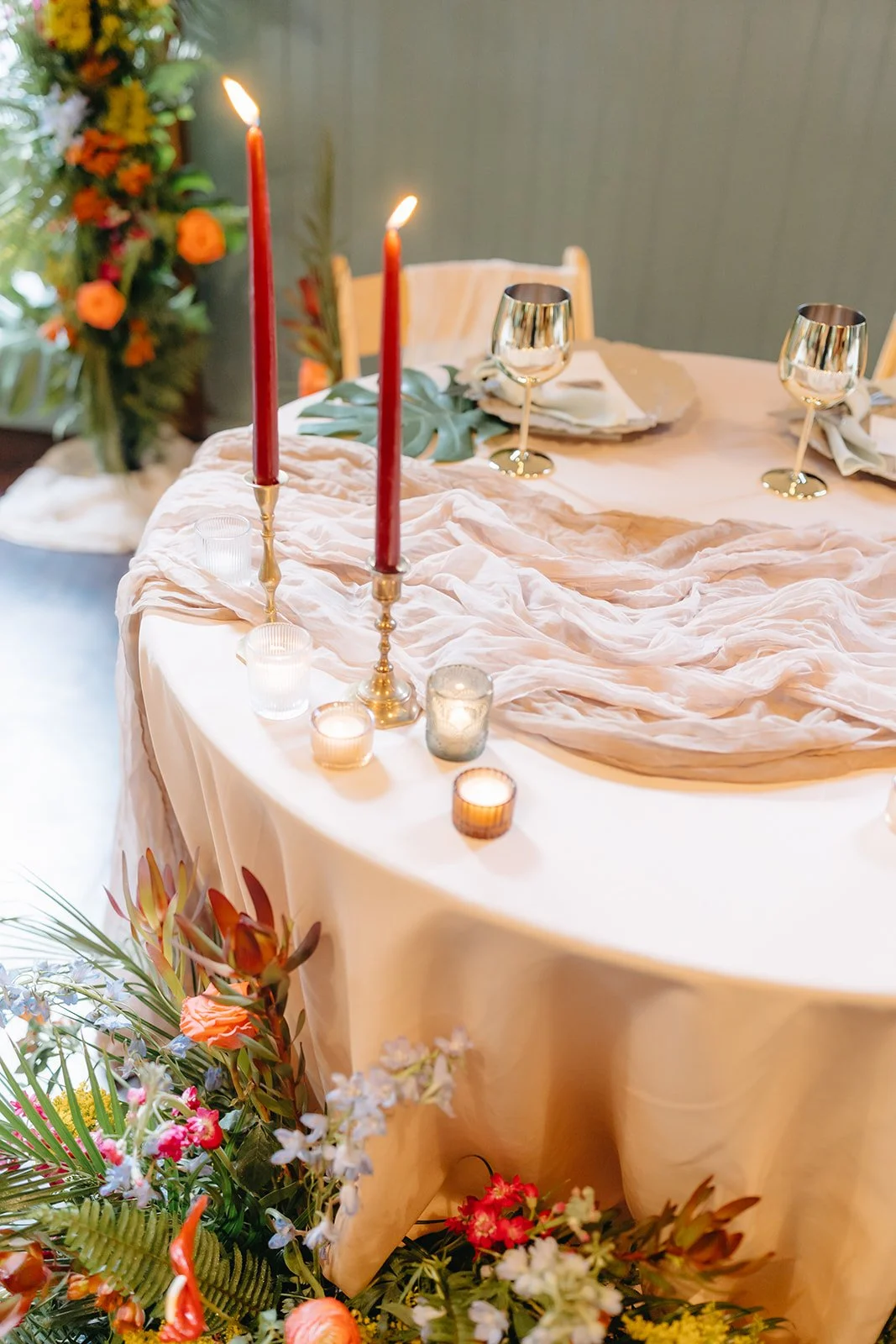 Elegant table setting with pink tablecloth, gold and metallic wine glasses, red and gold candles, votive candles, and floral arrangement in the background.
