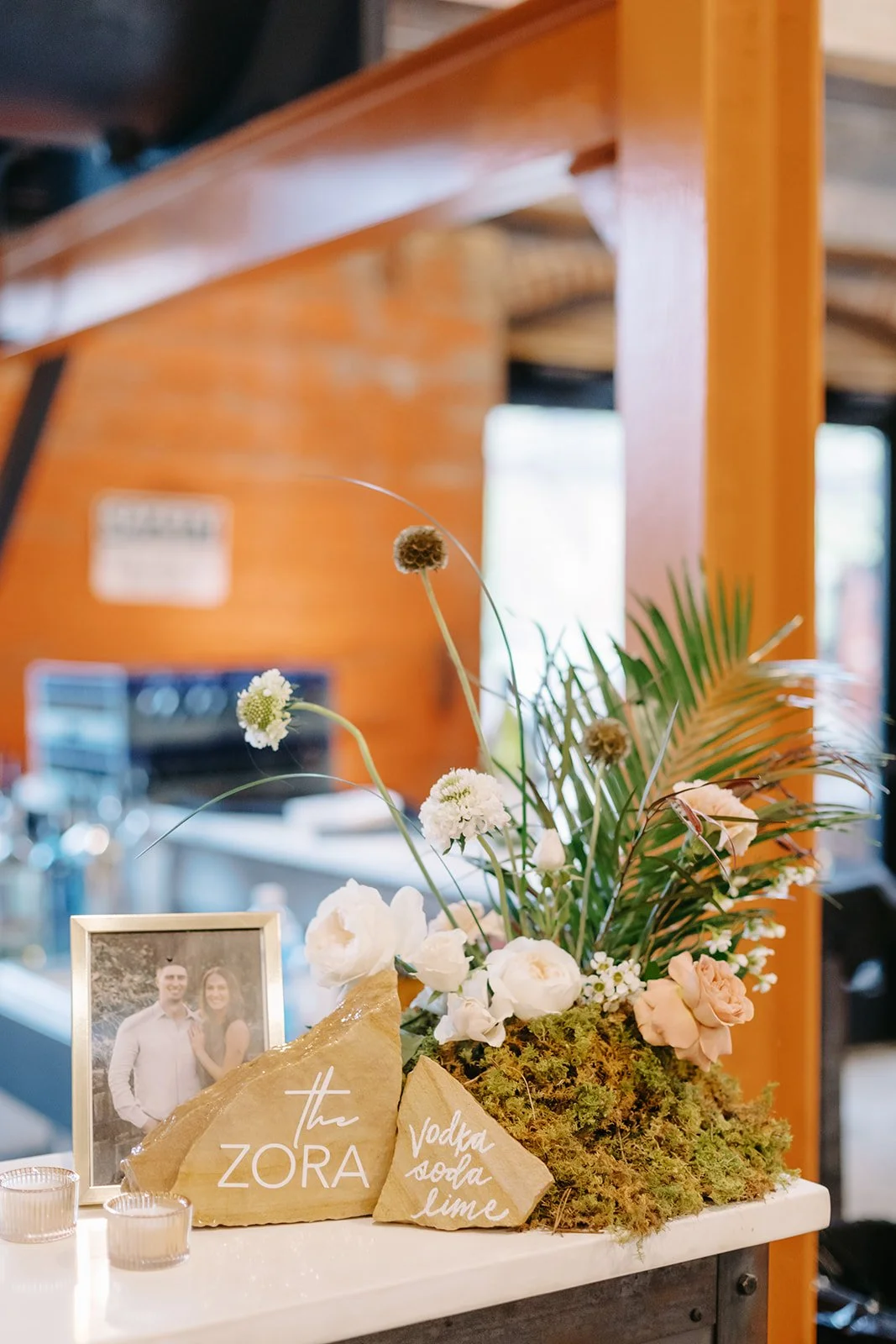 Decorative floral arrangement with white and peach flowers, greenery, moss, and beige stones with white lettering reading "the ZORA" and "vodka soda lime" on a white surface. A framed photo of a couple stands beside the arrangement.