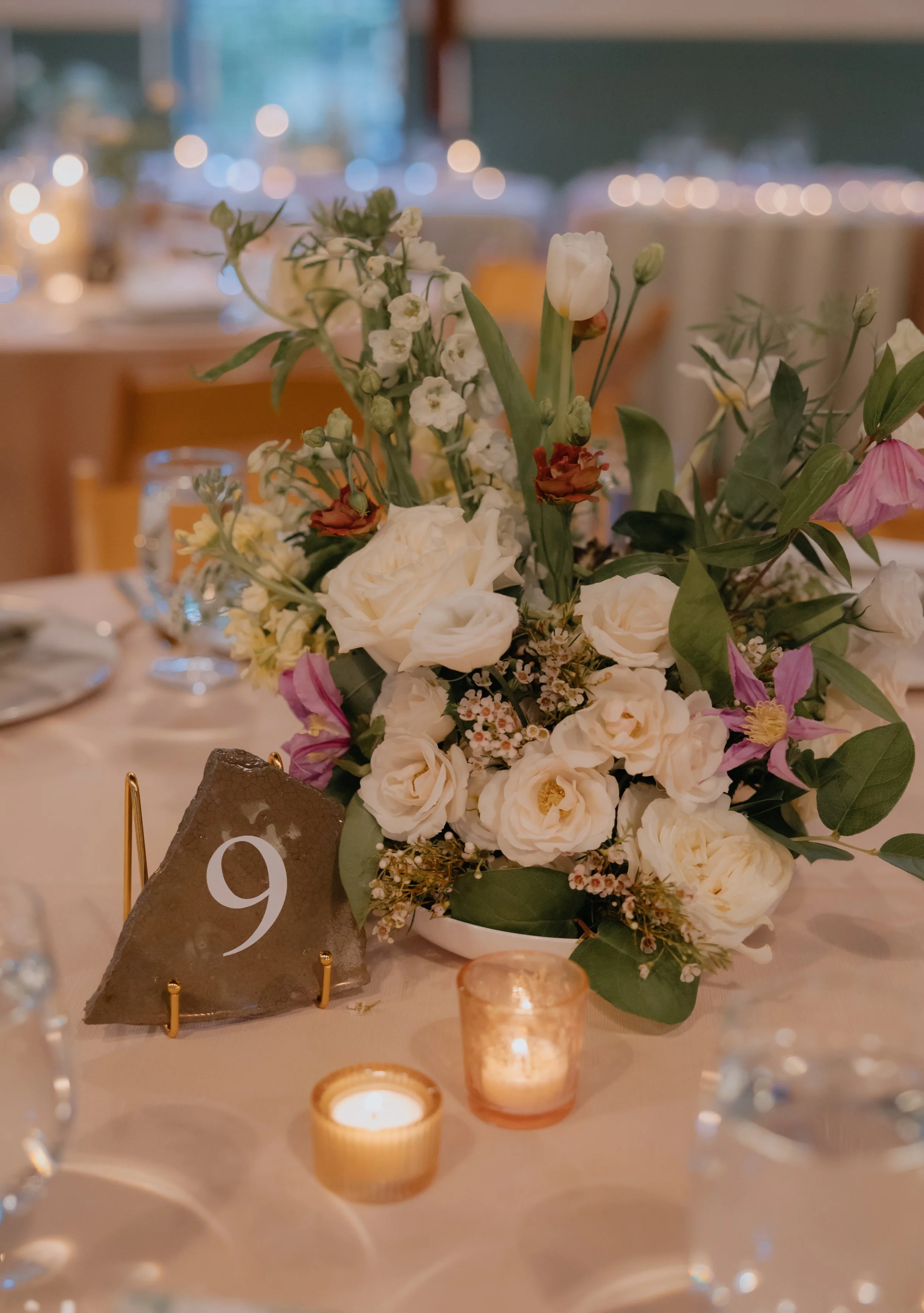 A floral centerpiece on a table with candles and a dark table number card that says '9'. The floral arrangement includes white roses, tulips, and other small white flowers with green leaves. The background is blurred with warm lighting.