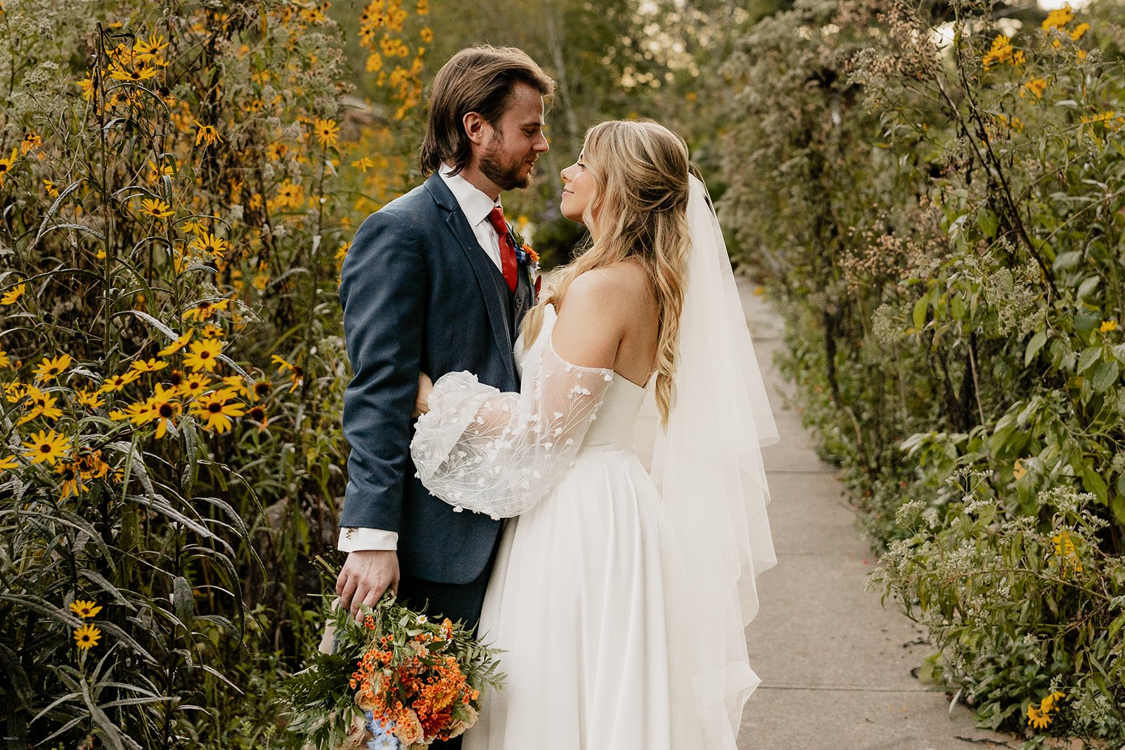 A bride and groom stand close together on a pathway surrounded by blooming yellow and purple flowers. The groom wears a dark blue suit with a red tie, and the bride wears a white wedding dress with off-the-shoulder lace sleeves and a flowing veil. They gaze at each other lovingly while holding a bouquet of orange and blue flowers.