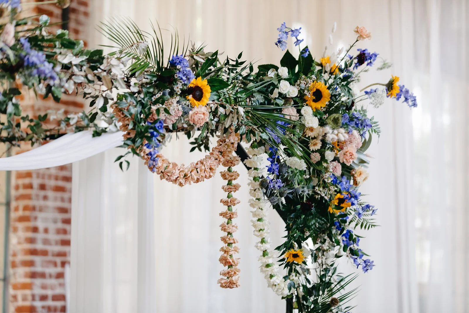 A floral arrangement with sunflowers, white roses, pink and white hanging garlands, and various green and purple foliage, set against a backdrop of brick and sheer curtains.