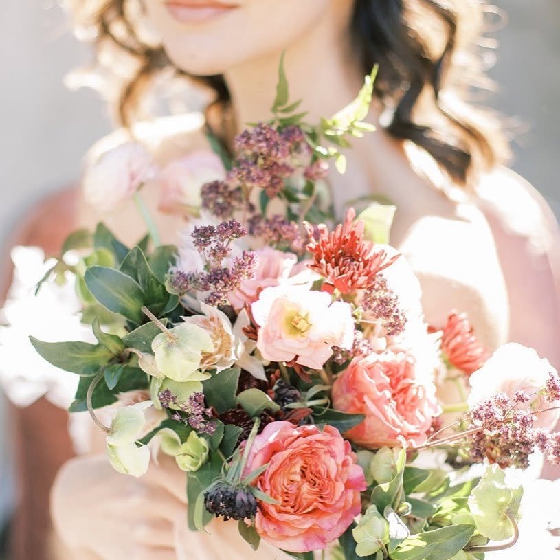 A woman in a light-colored dress holding a large bouquet of pink, peach, and purple flowers with green leaves.
