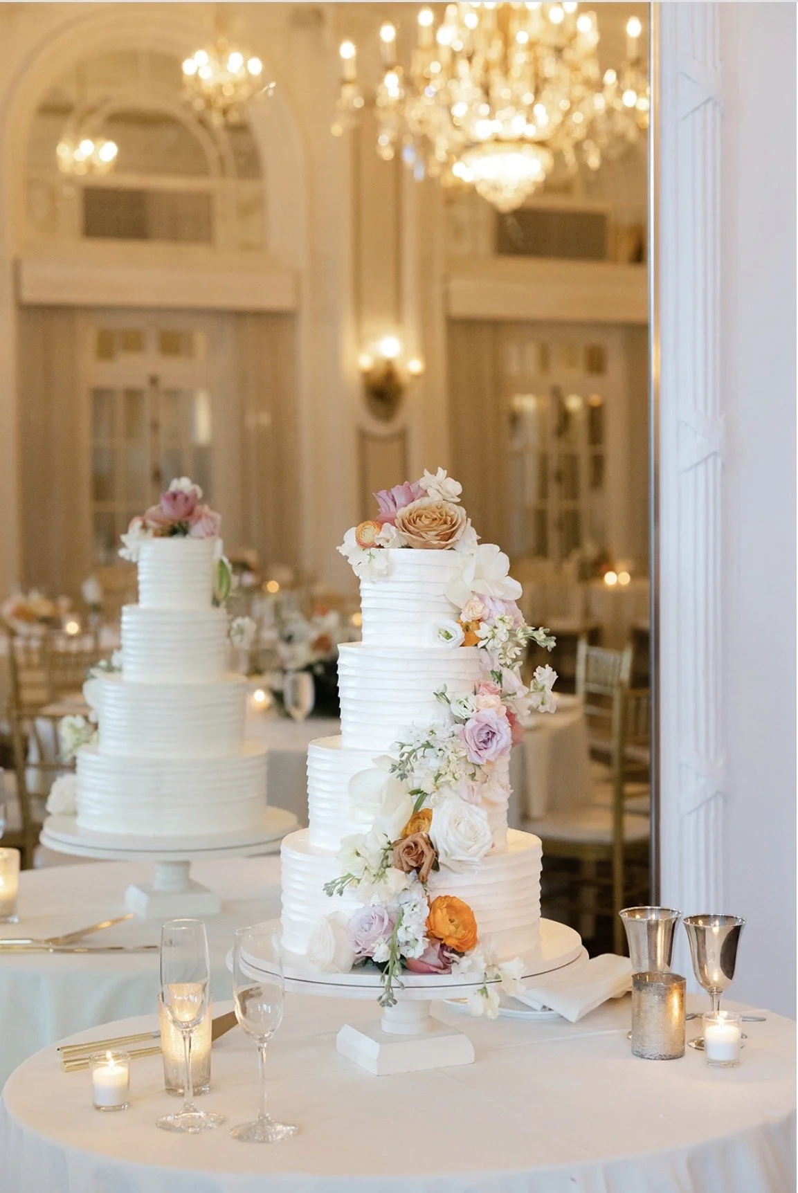 A multi-tiered wedding cake decorated with pastel-colored flowers on a round table, with candles and glassware, in an elegant banquet hall with chandeliers and tall windows in the background.