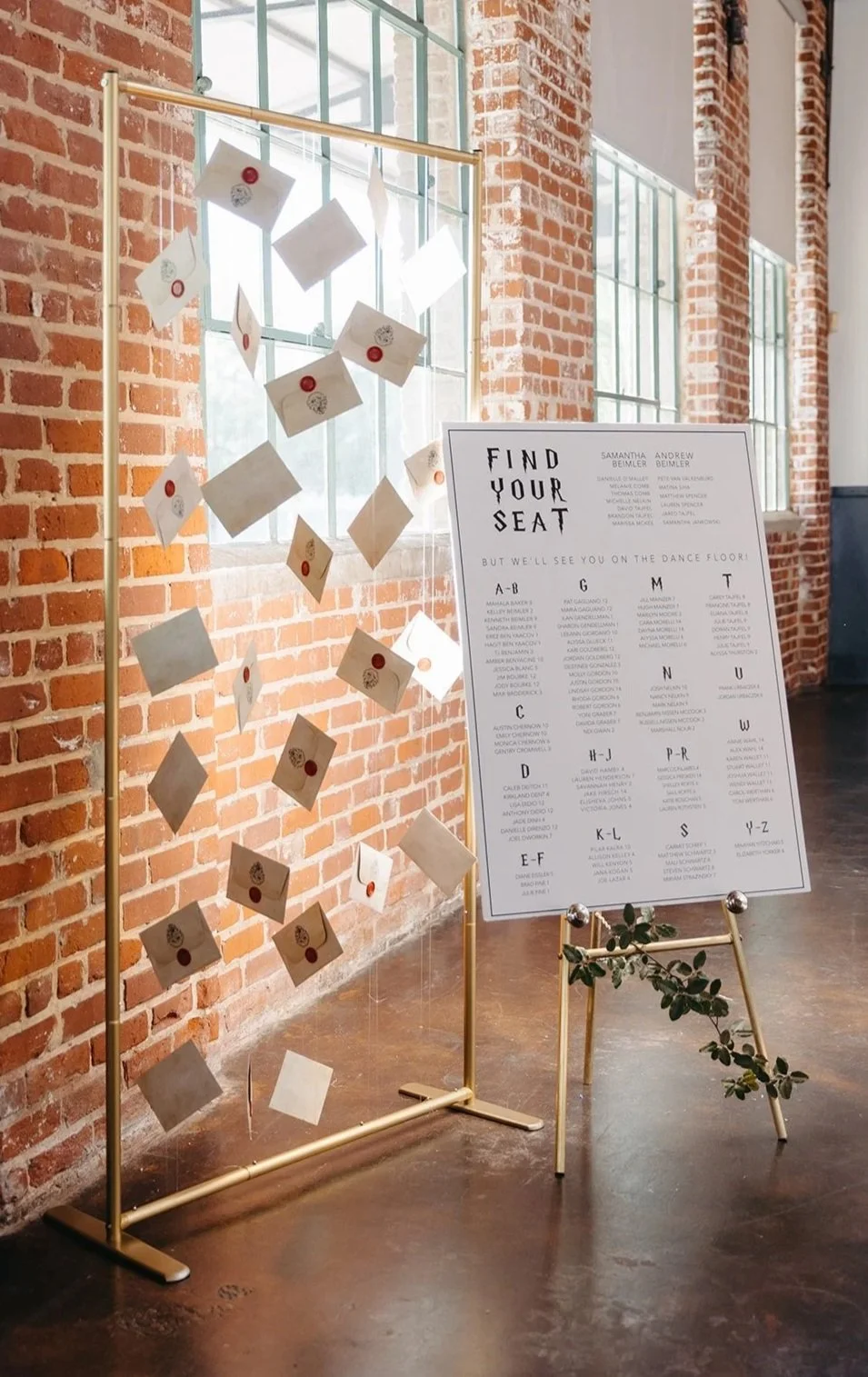 A wedding or event seating chart display with a gold stand, decorated with small envelopes and seals hanging in front of large windows and a red brick wall. Next to it, there's a sign on a stand with the title "Find Your Seat" and a list of names organized alphabetically by sections.