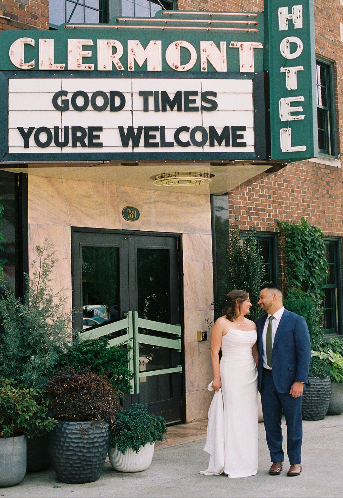 A bride and groom sharing a moment outside a building with a marquee sign that reads 'Clermont Hotel' and a message board that says 'Good Times You're Welcome.' The bride is in a white strapless wedding gown, and the groom is in a dark blue suit and a gray tie, standing close and smiling at each other.