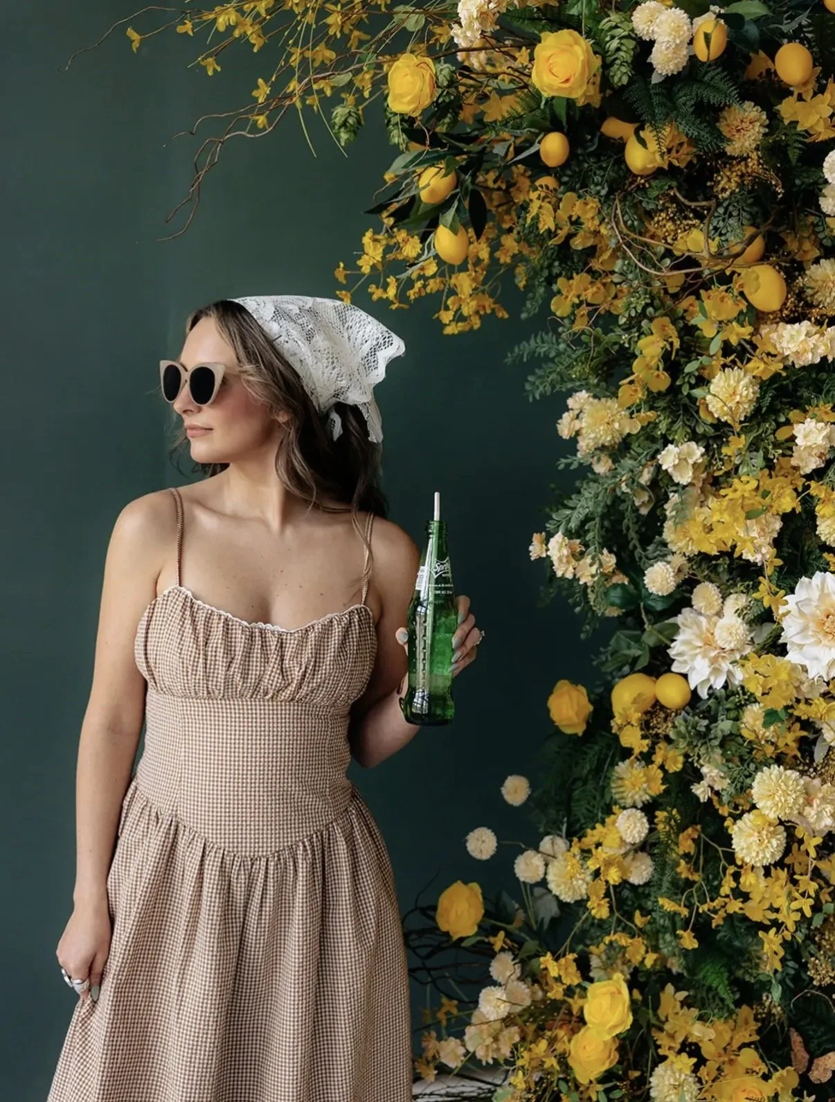 A woman wearing a gingham dress, sunglasses, and a lace headscarf holding a green glass soda bottle, standing next to a floral arrangement of yellow and white flowers against a dark green background.