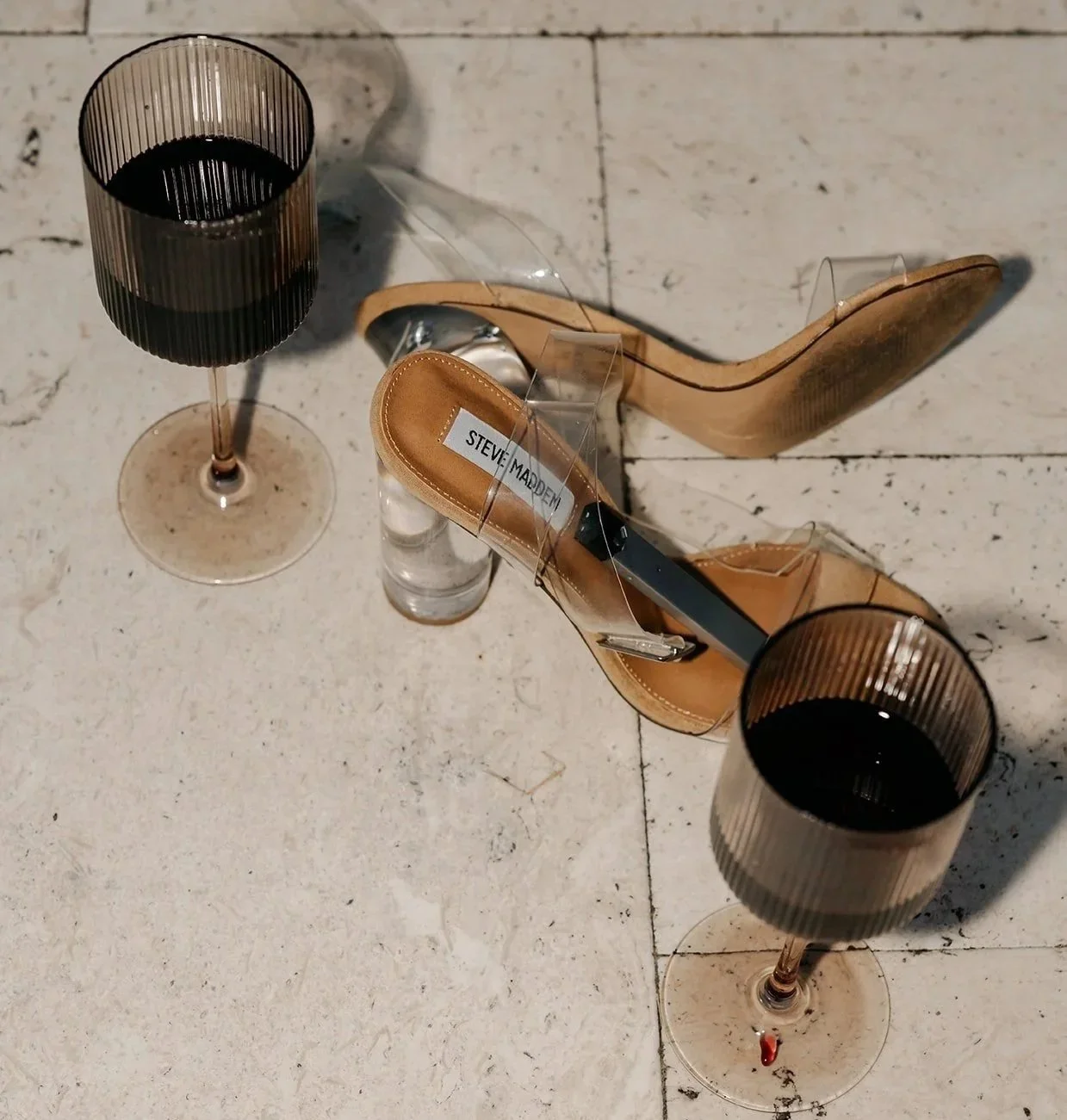 A pair of high-heeled sandals with a transparent strap, two glasses of red wine, and a bottle of water on a tiled floor.