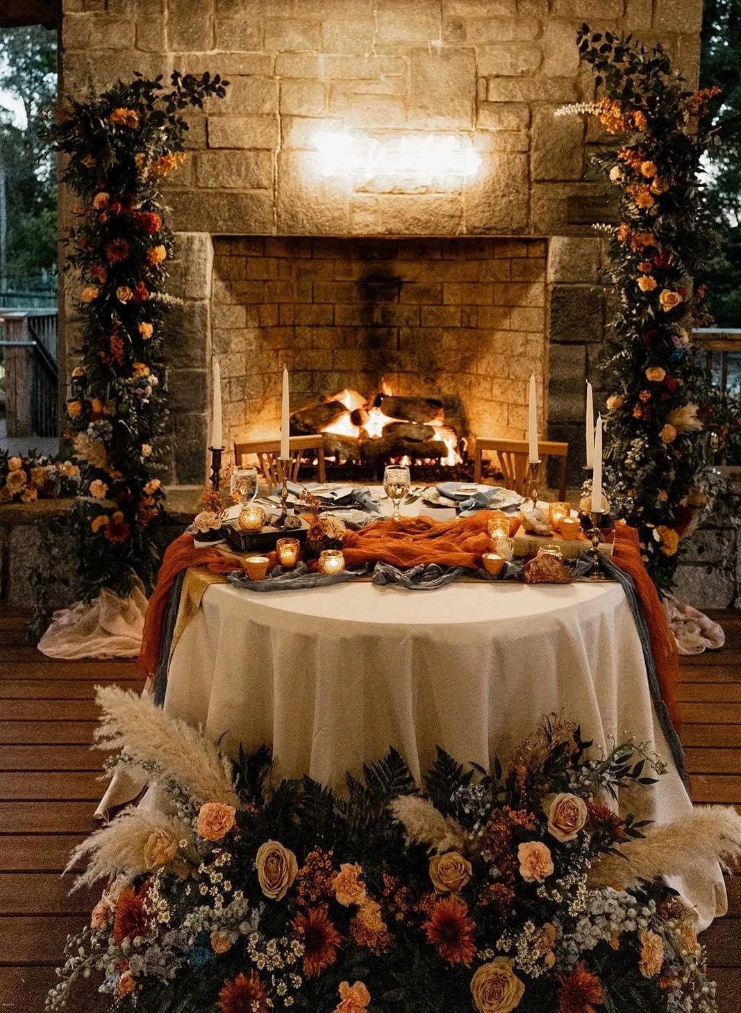 Wedding or special event setup with a round table covered with a white tablecloth, decorated with candles, flowers, and dark drapes, positioned in front of a stone fireplace with a fire burning inside, and floral arrangements decorating the fireplace and surrounding area.