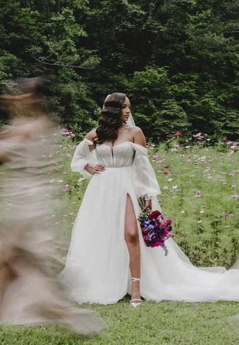 A woman in a white off-shoulder dress with a high slit stands in a garden with green grass and pink flowers, holding a colorful bouquet of flowers.