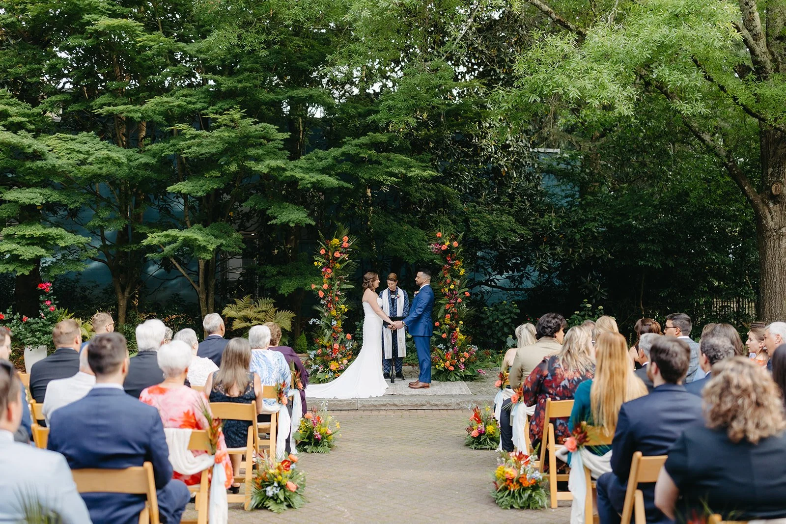 A wedding ceremony outdoors, with a couple holding hands and facing each other, surrounded by guests seated on wooden chairs, and decorated with colorful flowers and lush green trees in the background.