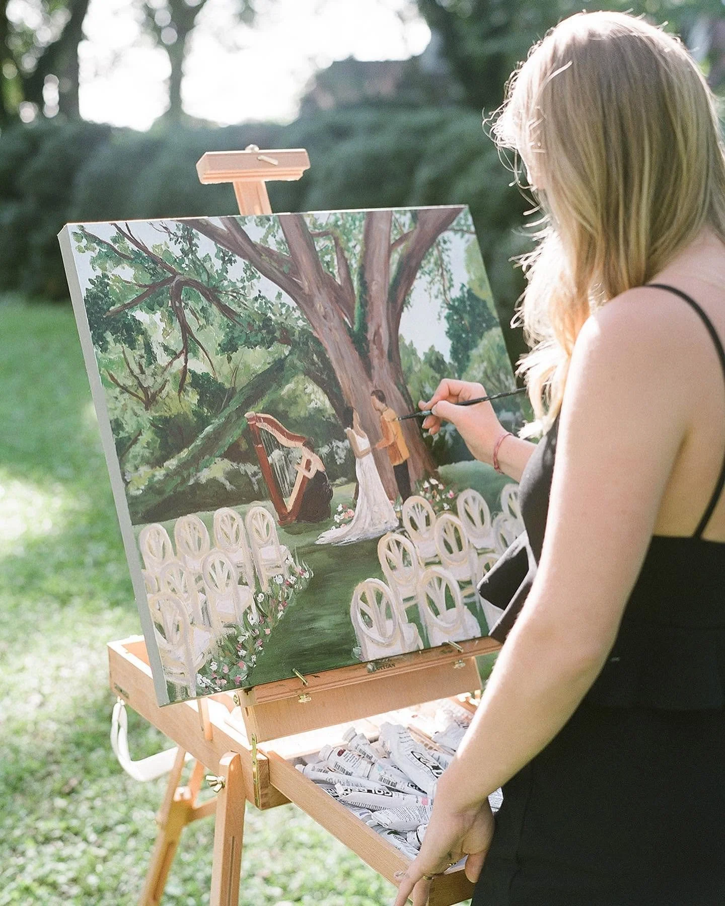 A woman with blond hair painting an outdoor wedding scene on a canvas, which is set up on an easel. The scene features chairs arranged as a ceremony, a large tree, and two figures in wedding attire.