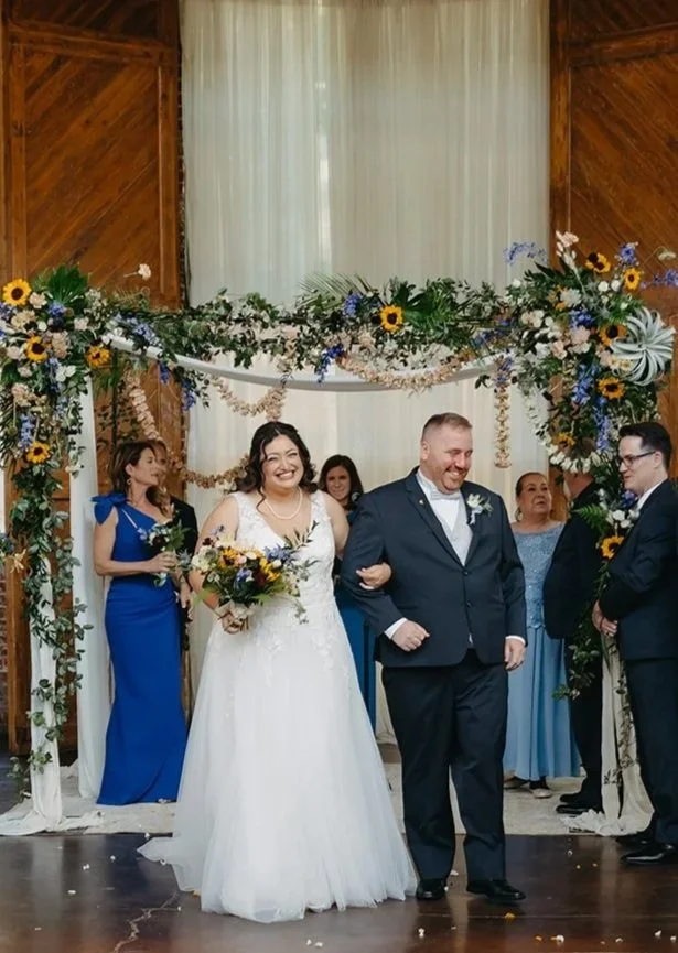 Bride and groom smiling, walking arm in arm under a decorated floral arch during a wedding ceremony. Female bridesmaids and guests stand in a rustic wooden venue.