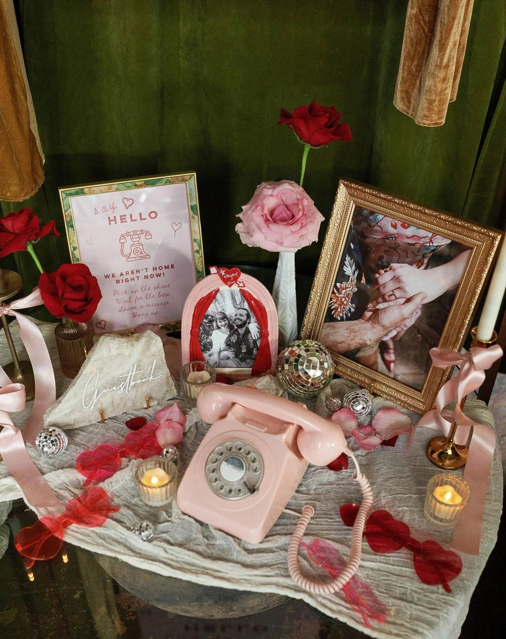 A decorative table display featuring a pink rotary phone, framed photos, candles, roses, heart-shaped decorations, a sign with a poem, and a bunch of pink rose petals scattered on the table. The background is green with curtains on the side.