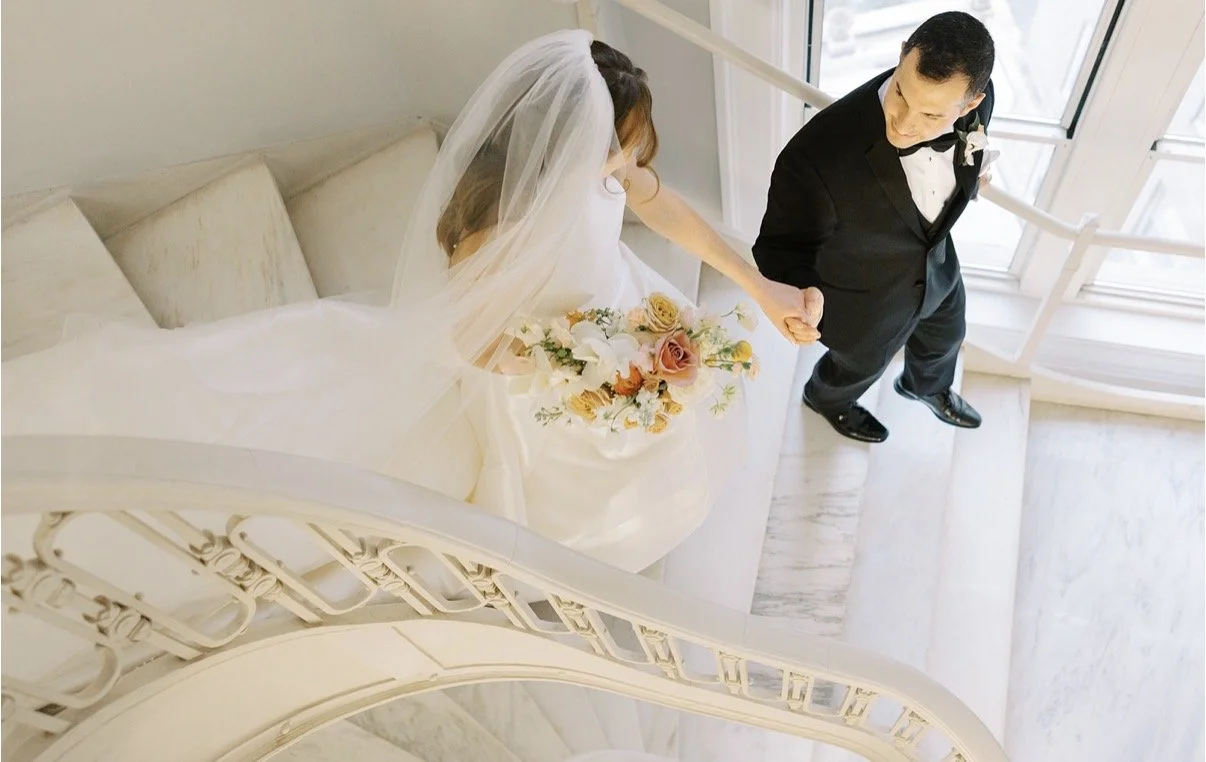 Bride and groom holding hands on a staircase, bride in a wedding dress and veil holding a bouquet of flowers, groom in a tuxedo, window in the background.