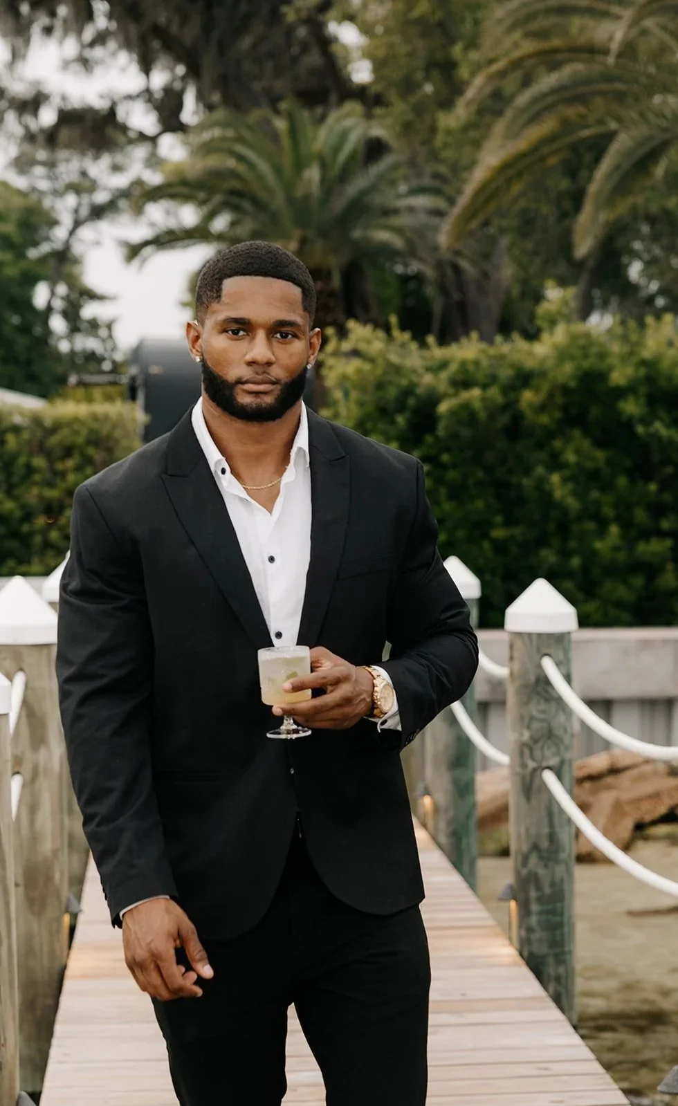 A young man in a black suit and white shirt, holding a cocktail, standing on a wooden pier with greenery and trees in the background.