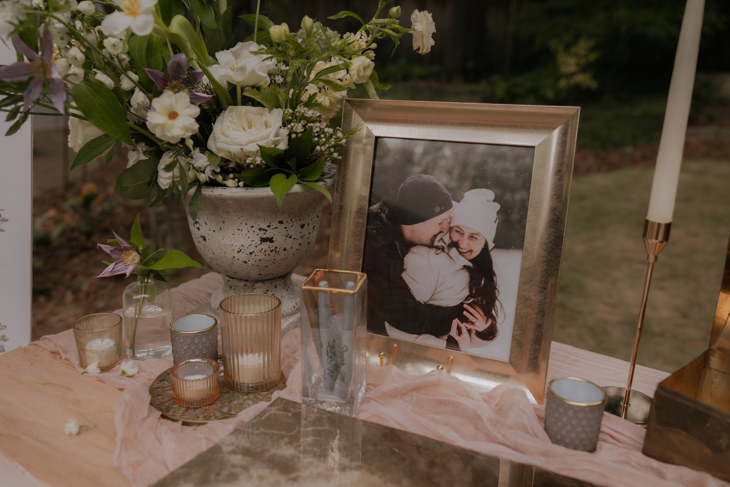 A memorial table with a large floral arrangement in a white speckled urn, a framed photograph of a smiling couple in winter clothing, and small decorative candles on a pink cloth.