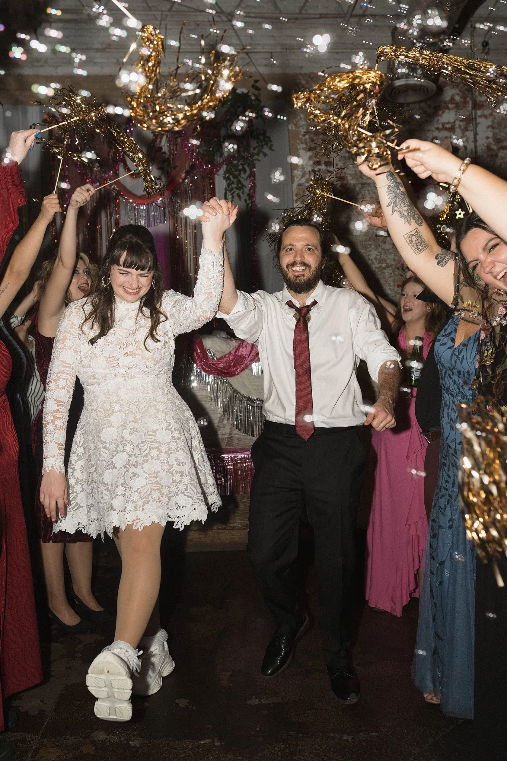 A group of people celebrating at a party, holding gold and black tinsel and sticks high in the air. The woman in front is wearing a white lace dress and sneakers, smiling with eyes closed, dancing. The man next to her is in a white shirt with rolled-up sleeves, dark pants, and a red tie, smiling and holding hands with her. Other partygoers in colorful dresses and outfits are smiling and enjoying the celebration, with decorative streamers and tinsel hanging from the ceiling in the background.