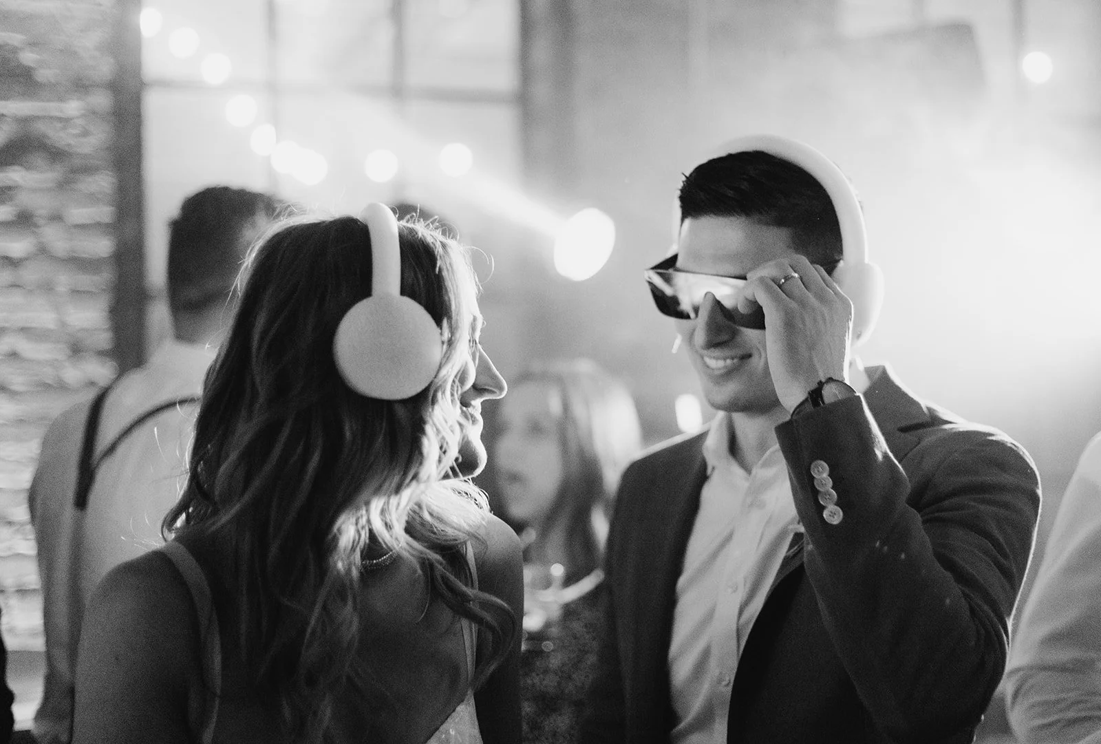 Black and white photo of a man and woman dancing at a party, both wearing headphones. The man is smiling and adjusting his headphones, the woman is looking at him, smiling. People are blurred in the background.