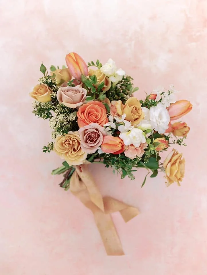 A bouquet of pastel-colored roses, tulips, and white flowers tied with a beige ribbon on a pink background.