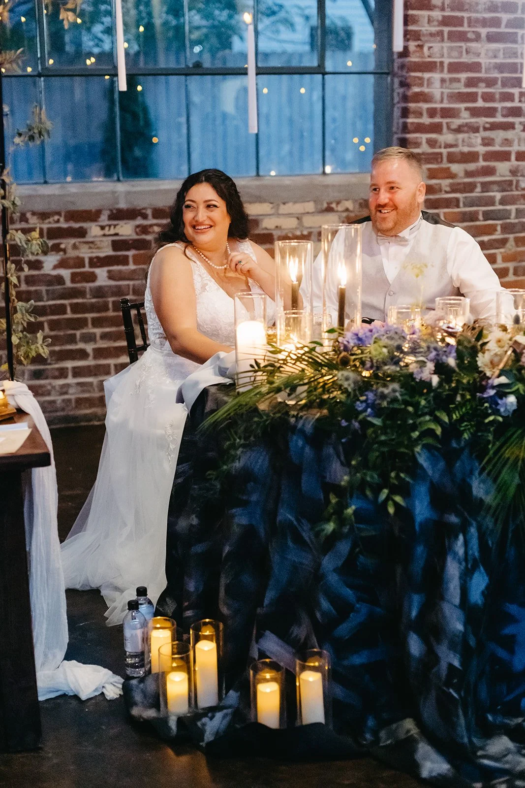A bride and groom sitting at a table during their wedding reception, smiling and looking at each other, with candles and floral arrangements on the table, in a rustic indoor venue with exposed brick walls and large windows.