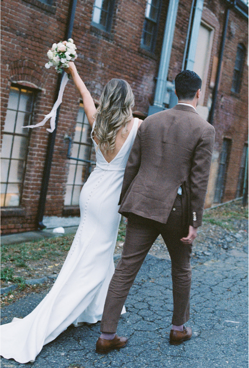 Bride in a white wedding gown holding a bouquet with her arm raised, standing next to a groom in a brown suit, on an outdoor cobblestone street in front of a brick building.