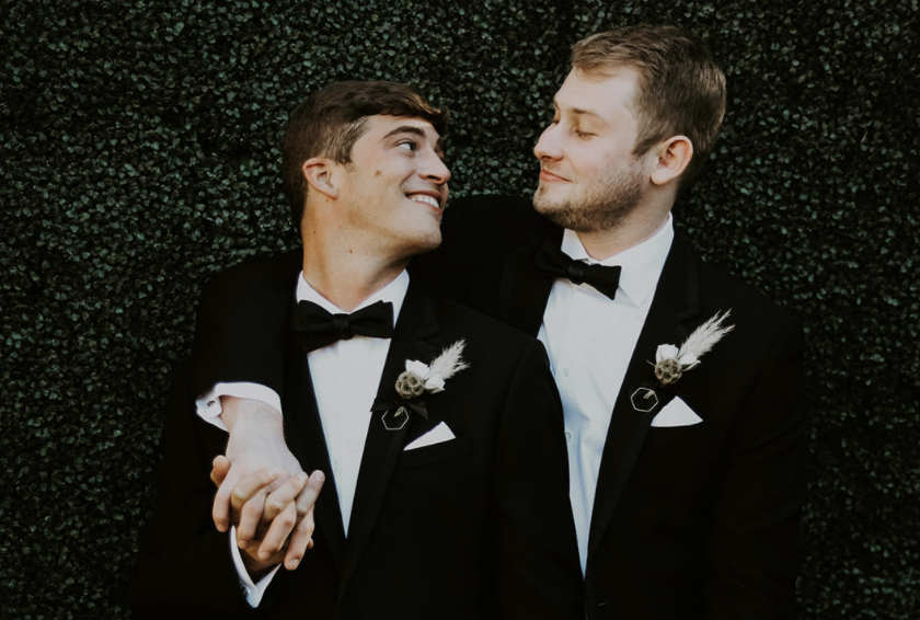 Two men in tuxedos and bow ties smiling at each other, holding hands, against a green foliage background.
