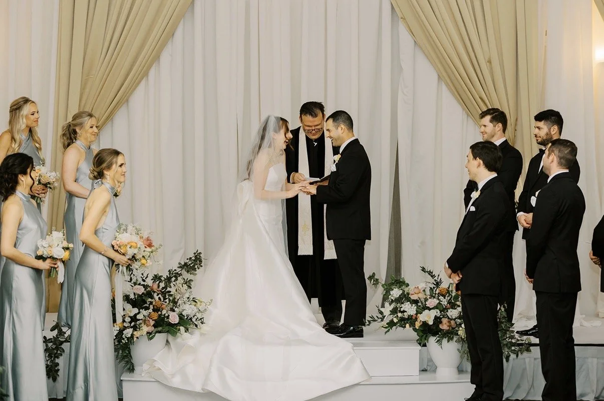 Wedding ceremony with a bride and groom exchanging rings, surrounded by bridesmaids and groomsmen, decorated with flowers and drapes.