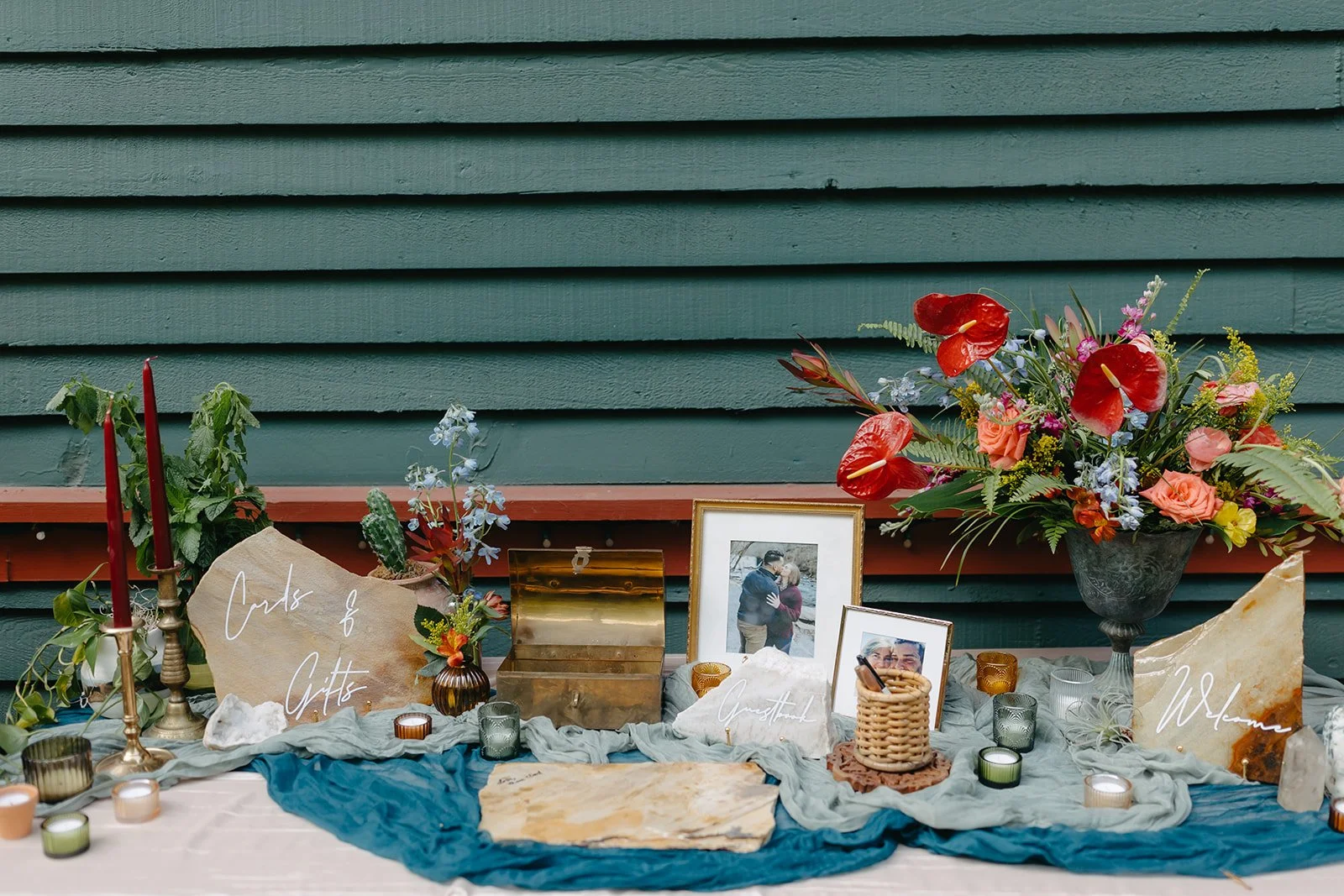 A memorial display with candles, framed photographs, flowers, and decorative signs on a table against a green wooden wall. Signs read 'Cards & Gifts' and 'Welcome'.
