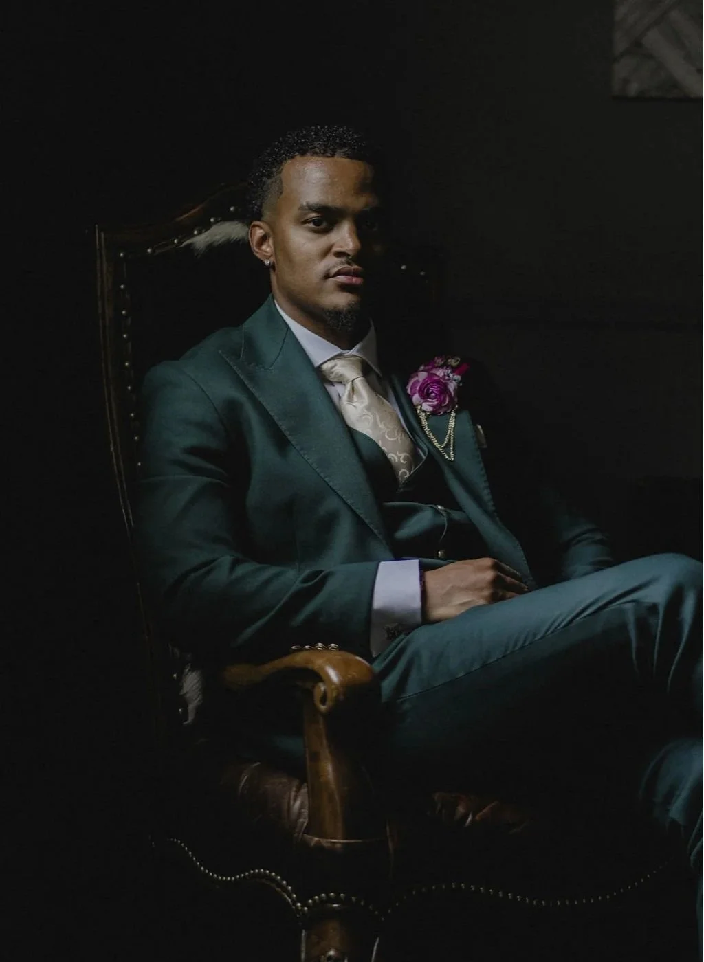 A man in a dark green suit and tie sitting on a wooden chair with a high back, in a dimly lit room.