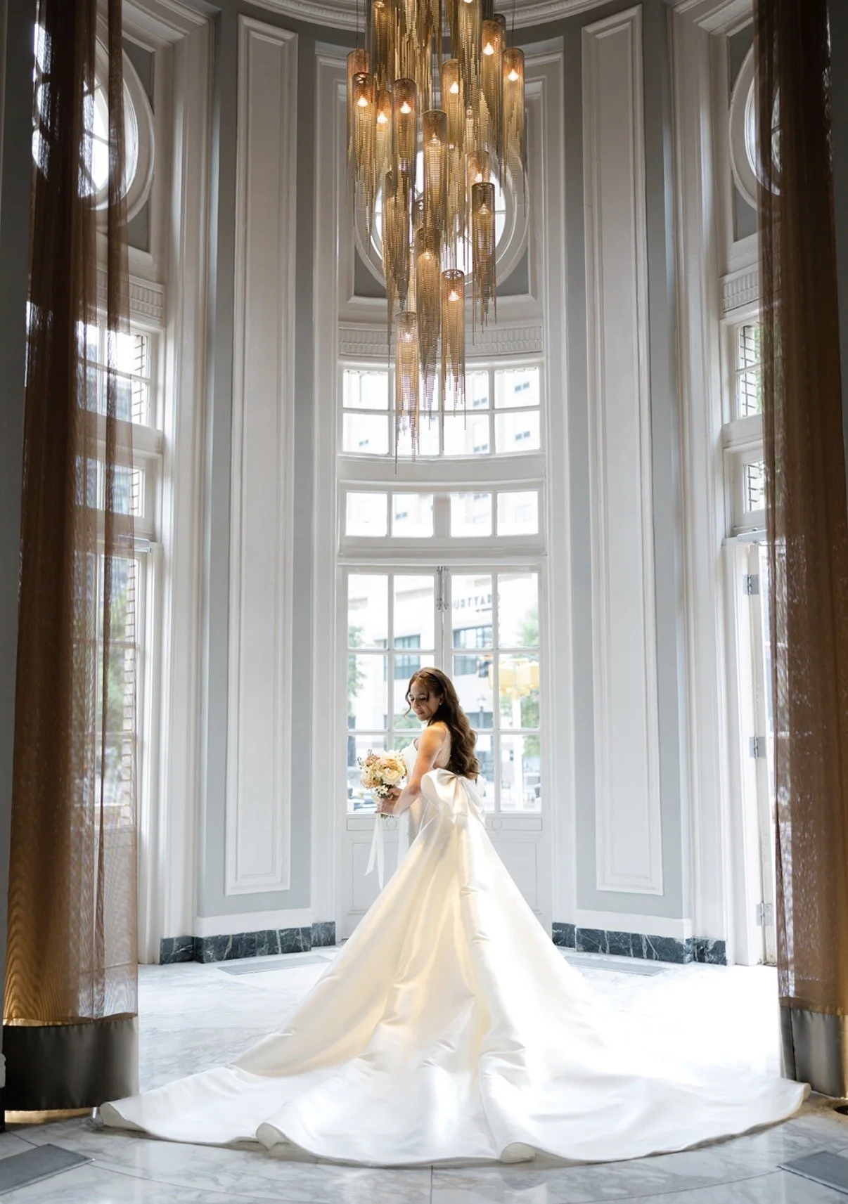 Bride in a white wedding gown holding a bouquet of flowers, standing in front of large windows in a bright, elegant room with a modern chandelier hanging from the ceiling.