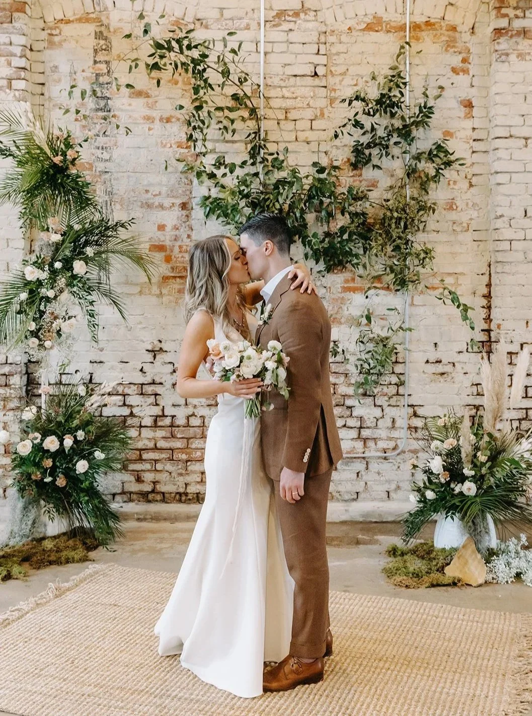 A bride and groom sharing a kiss during their wedding ceremony, standing in front of a rustic brick wall decorated with green foliage and white flowers, with a woven rug and floral arrangements on the ground.