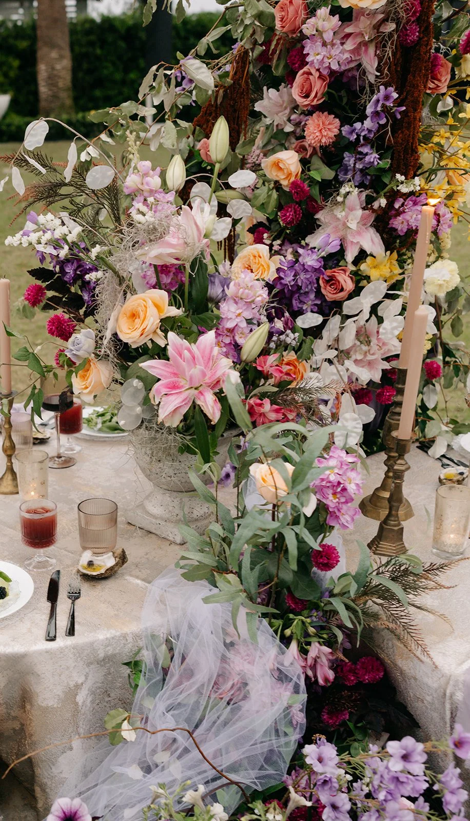 A large floral centerpiece with pink lilies, roses, and purple, white, and pink flowers on a decorated table at an outdoor event.