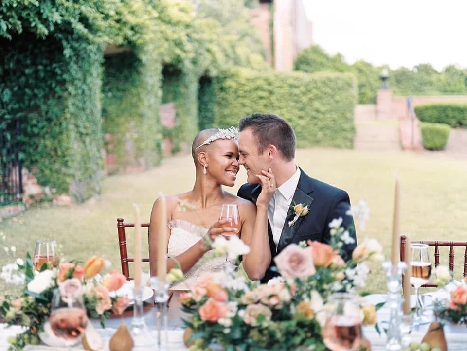 A newlywed couple sits at a decorated outdoor reception table, smiling and touching foreheads with their noses close together, surrounded by flowers and drinks.