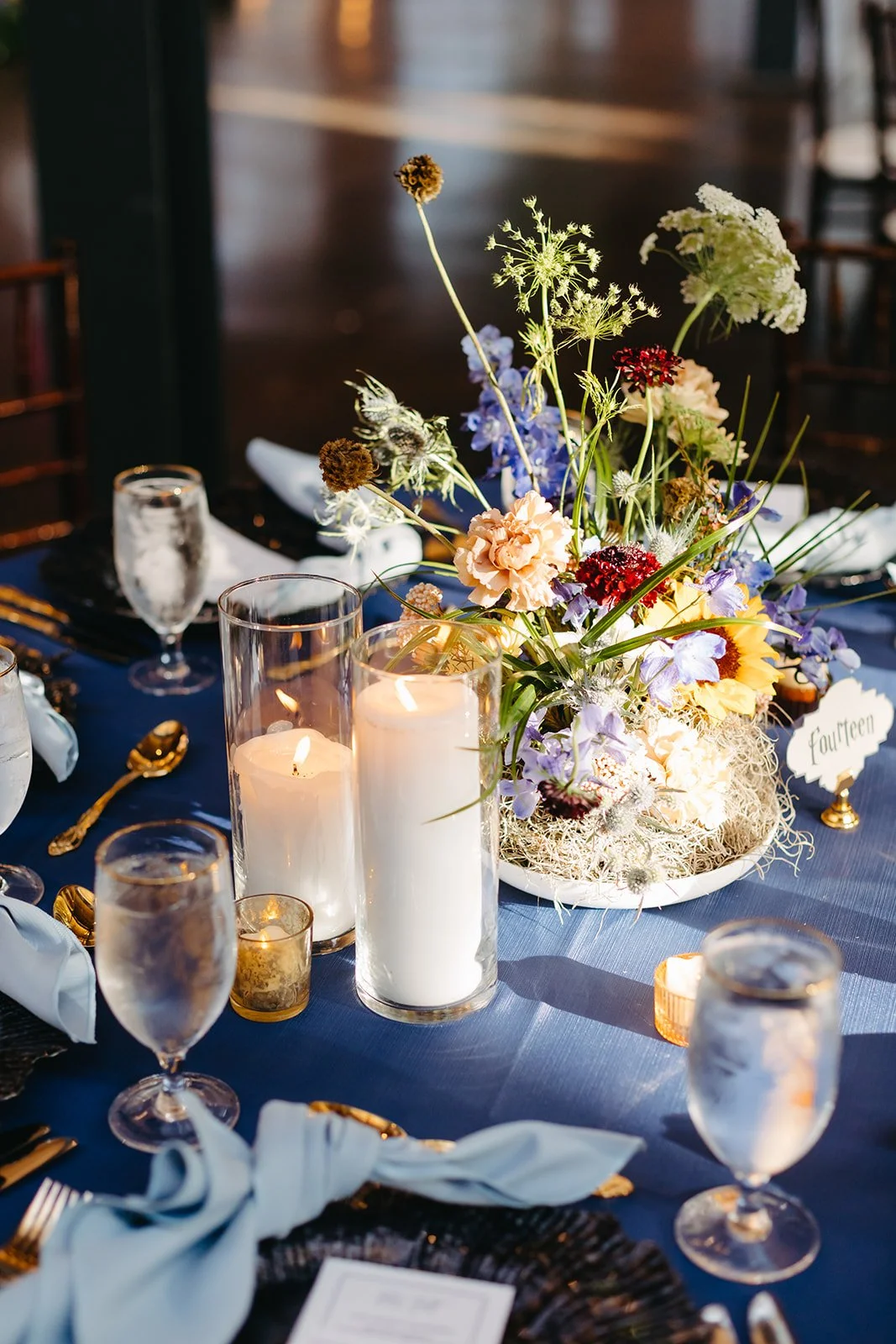 Elegant table arrangement with a floral centerpiece, candles, wine glasses, and a table number card on a navy tablecloth.