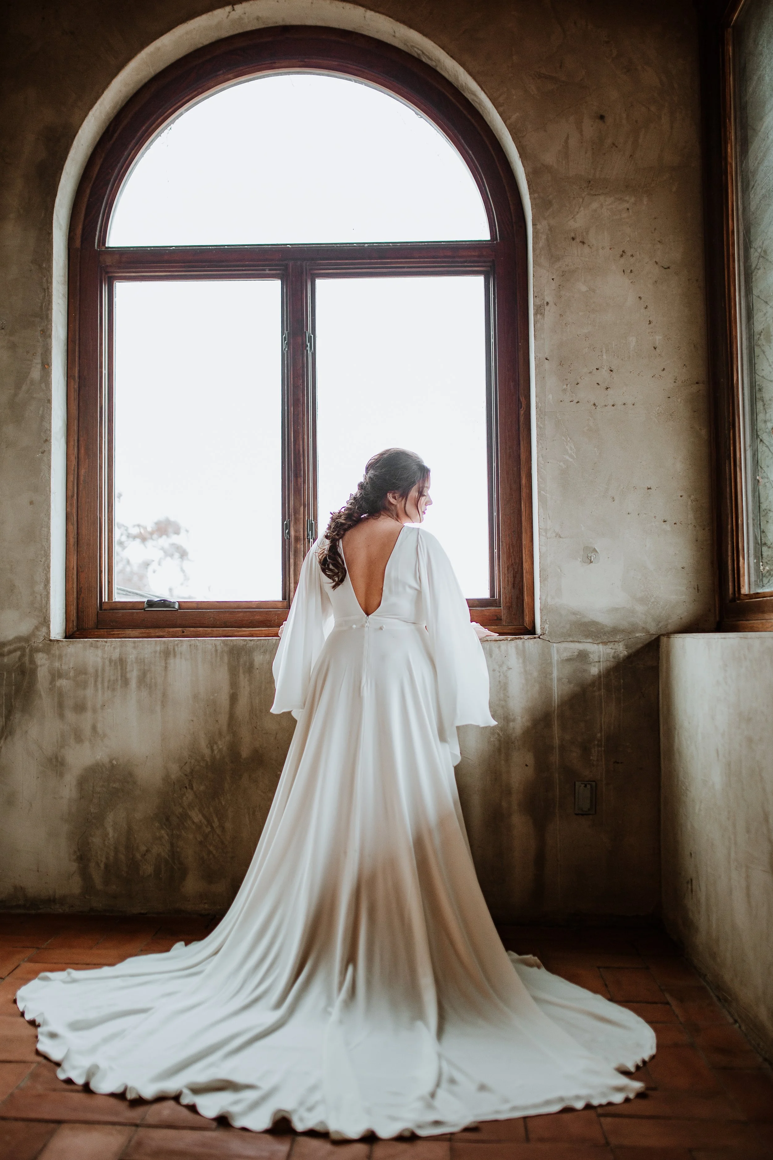 A bride in a white wedding gown with a long train standing indoors in front of a large arched window, with her back facing the camera.