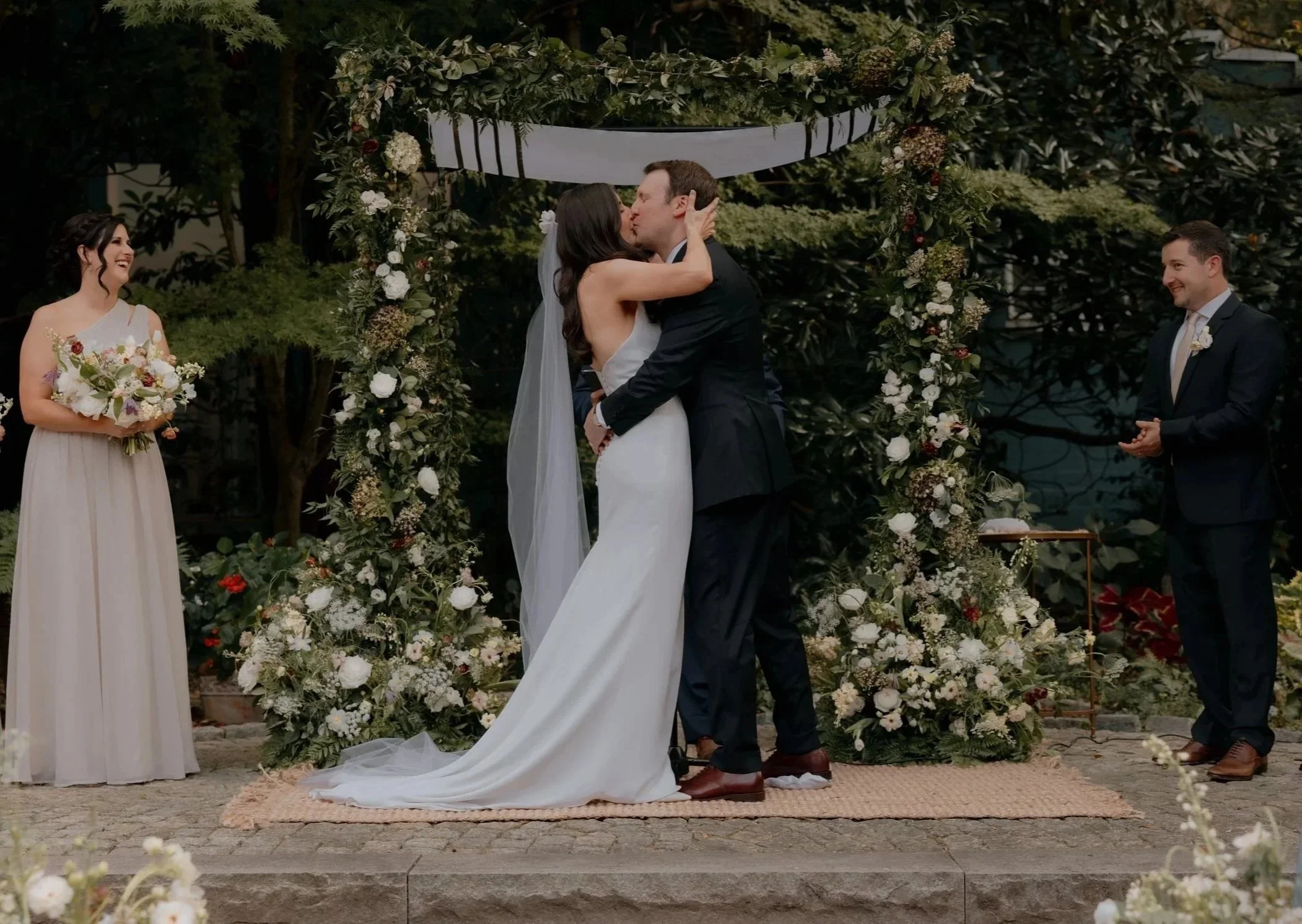 A wedding ceremony with a couple kissing at an outdoor altar decorated with flowers. The bride is wearing a white wedding dress and the groom a dark suit. Two women and one man are standing nearby, smiling, with a lush green background.
