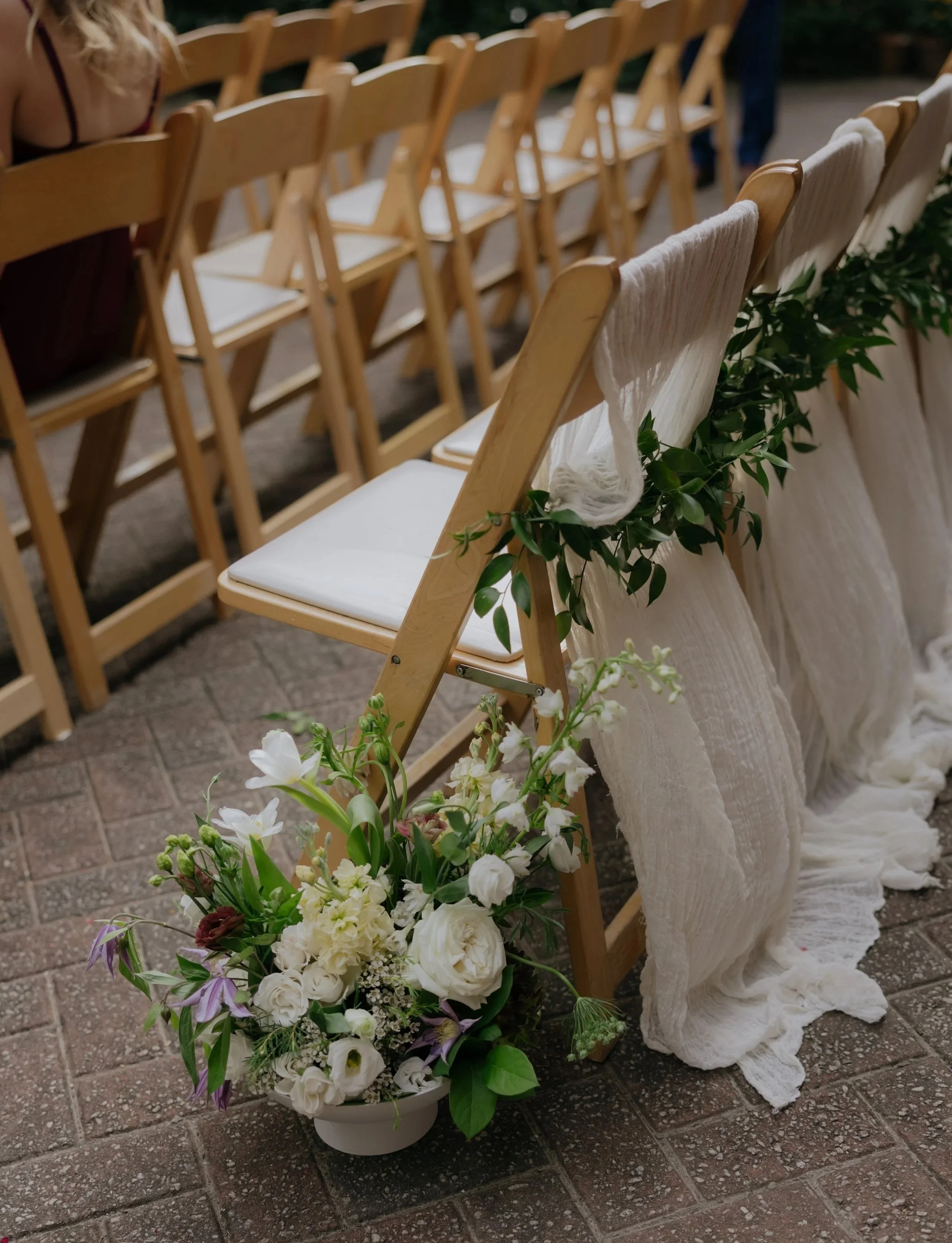 Rows of wooden chairs decorated with white cloth and green foliage, with a large floral arrangement of white and purple flowers on the ground, likely part of a wedding or similar ceremony setup.