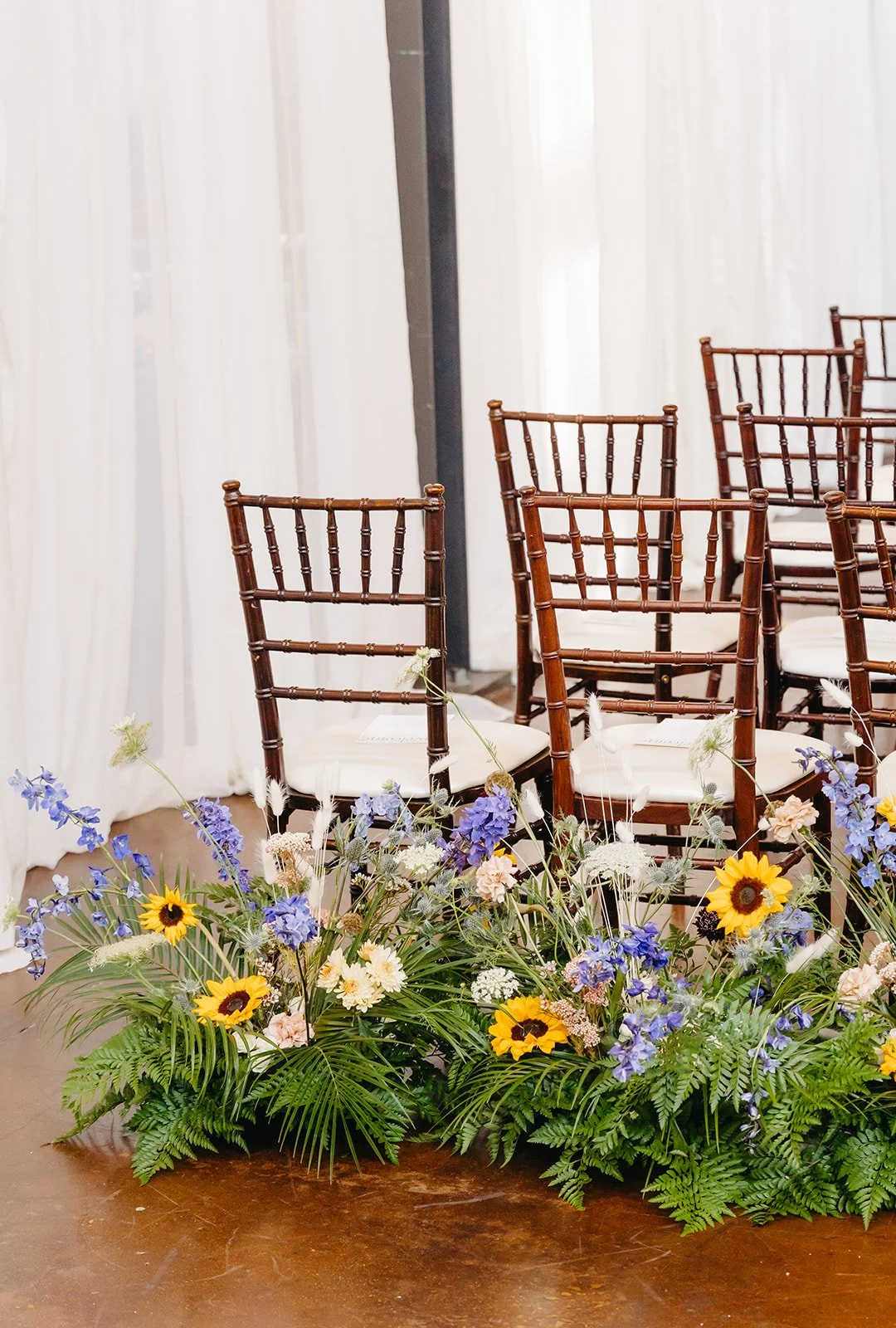 Rows of wooden chairs arranged for a wedding ceremony in front of a floral arrangement with yellow sunflowers, purple and white flowers, and green foliage.