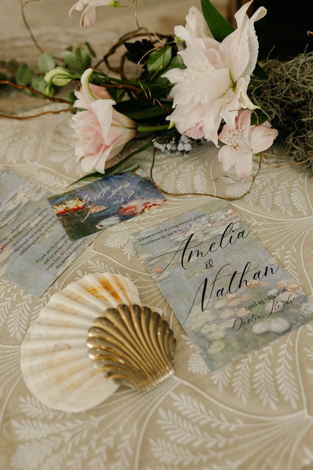 Wedding invitation card with watercolor landscape background, featuring the names Amelia and Nathan, and date April 23-25, 2026, placed on a table with a floral arrangement including pink and white lilies, seashell, and leaf-patterned tablecloth.