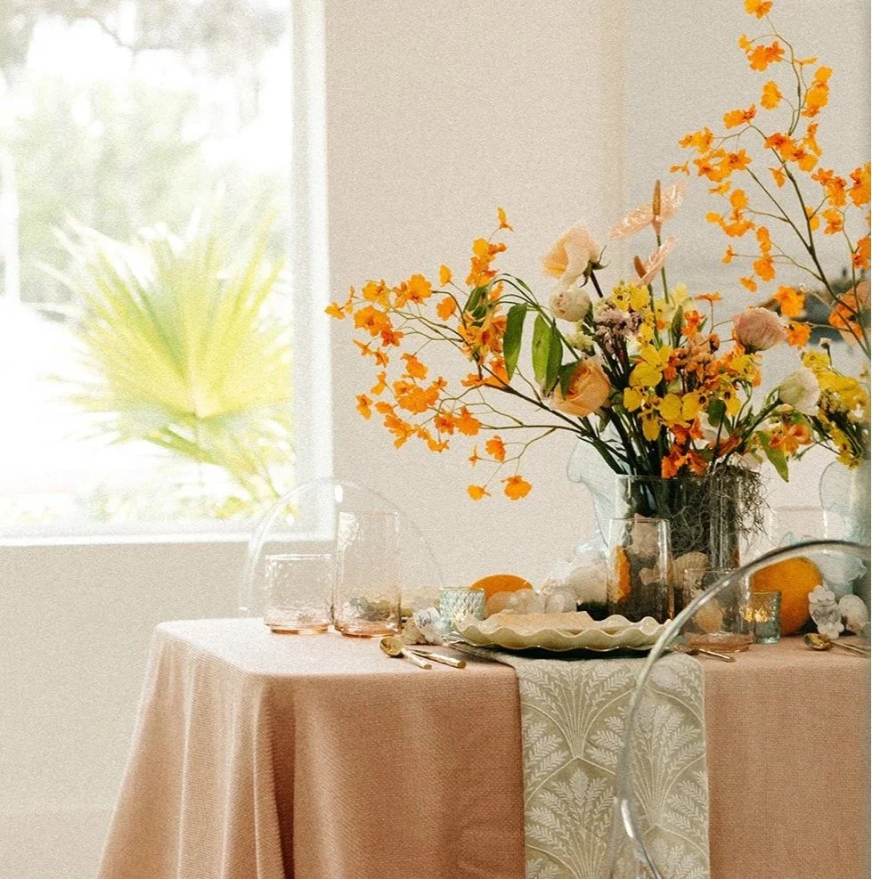 Decorative dining table with a large floral centerpiece in glass vases, a tray with small objects, a table runner, gold utensils, and glassware, set in a bright room with a window and a view of green plants outside.