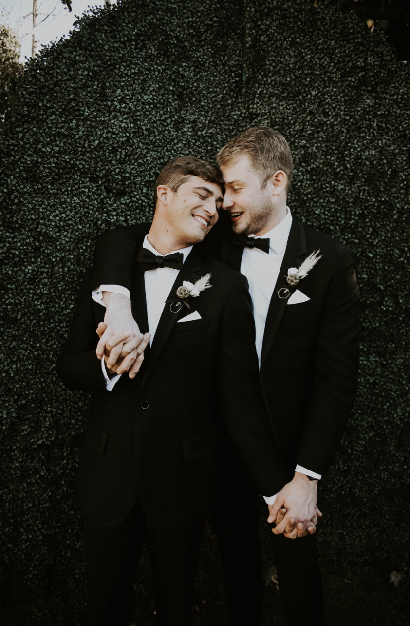 Two men in tuxedos holding hands, smiling and leaning their heads together in front of a green hedge.