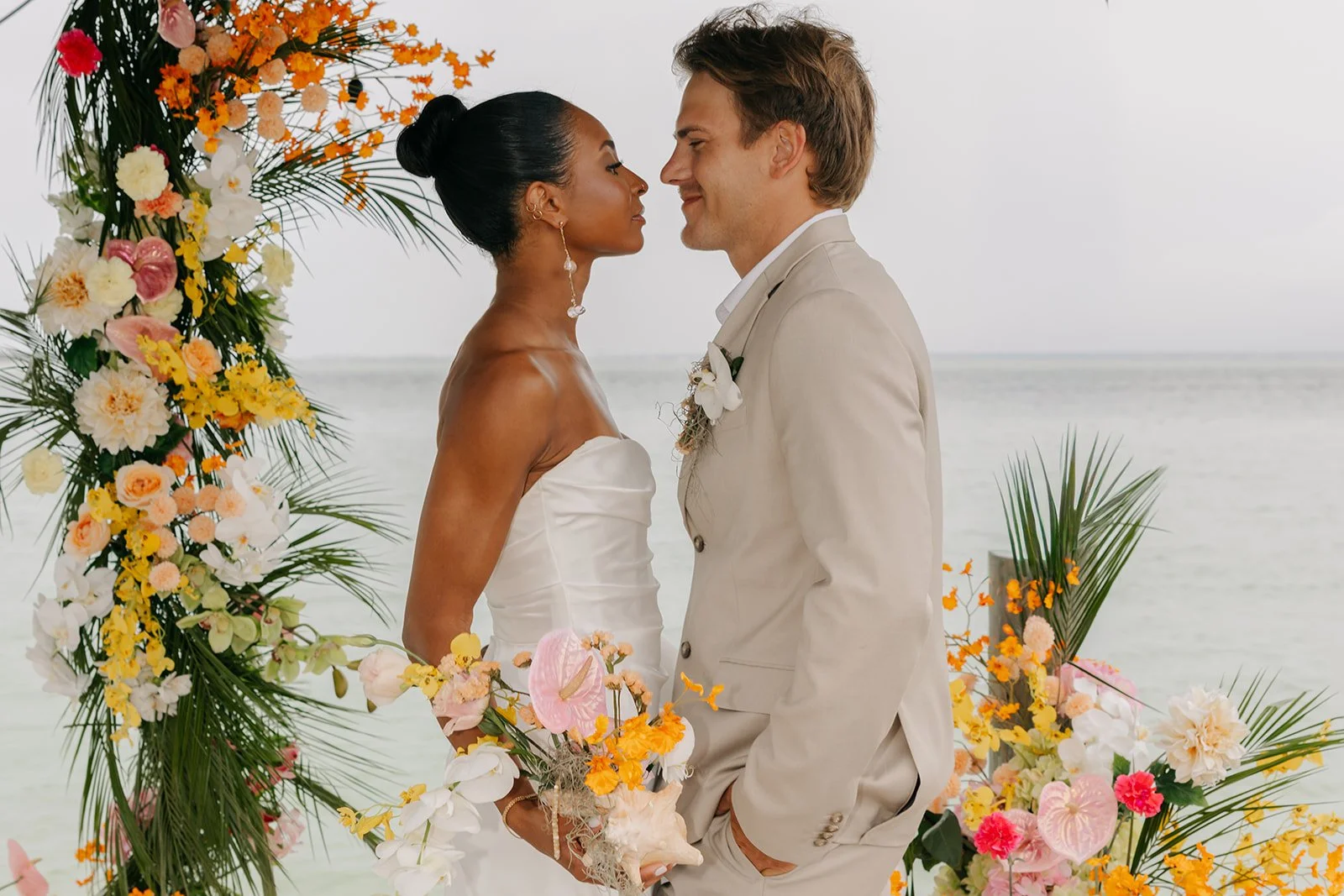 A couple getting married on the beach, standing close to each other surrounded by colorful tropical flowers and greenery, with the ocean in the background.