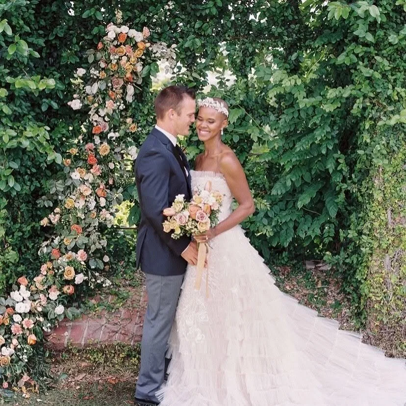 A couple dressed in wedding attire, standing close together and smiling, in front of a floral arch and lush green foliage, during a wedding ceremony.