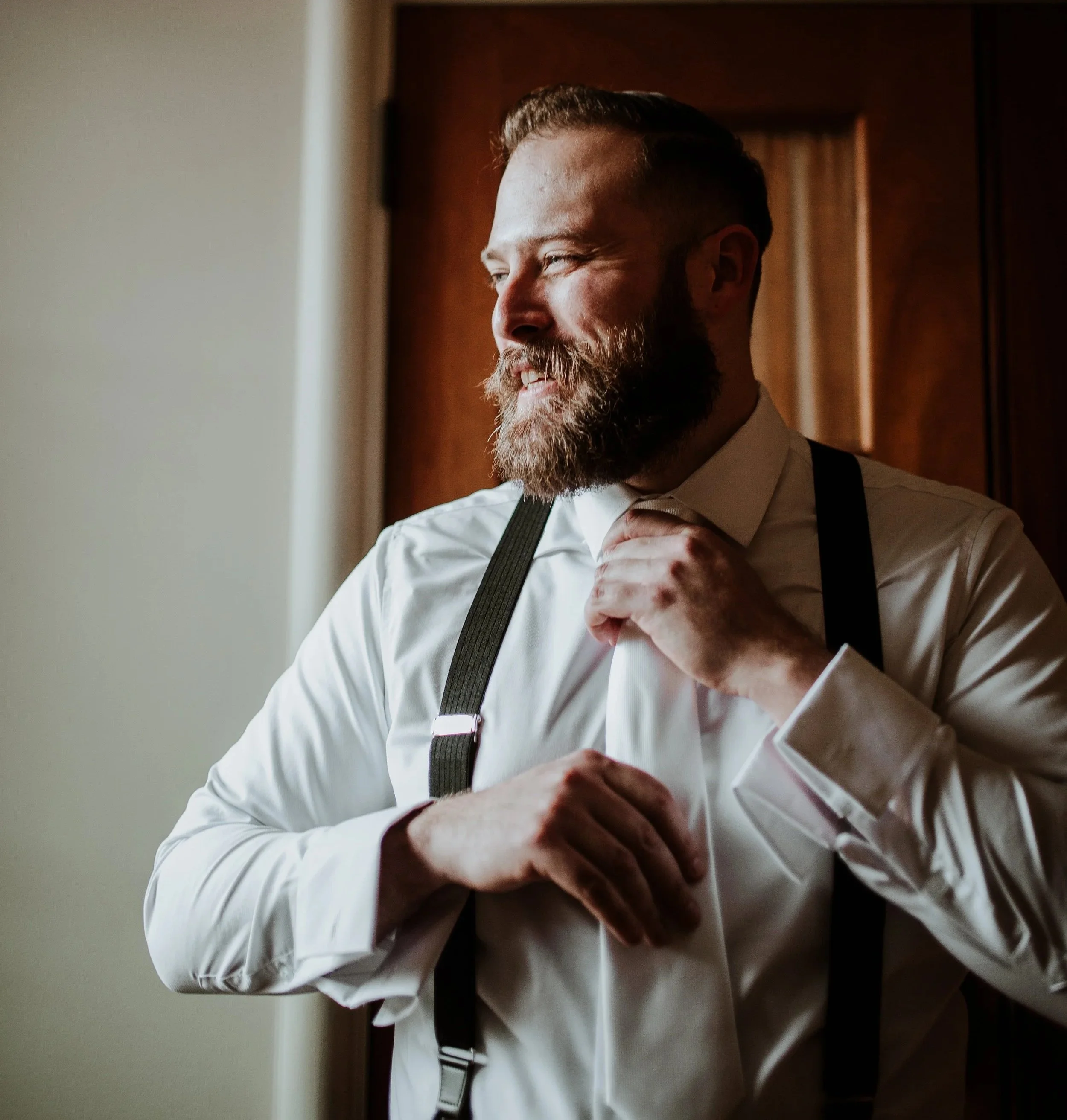 A man with a beard and short hair, dressed in a light-colored dress shirt and dark suspenders, adjusting his collar and tie while smiling near a window with a wooden door in the background.