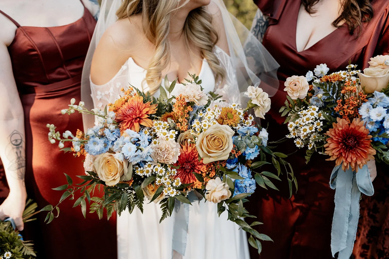 A bride in a white wedding dress holding a bouquet of various flowers including roses, daisies, and chrysanthemums in shades of cream, orange, and blue, surrounded by bridesmaids in burgundy dresses.