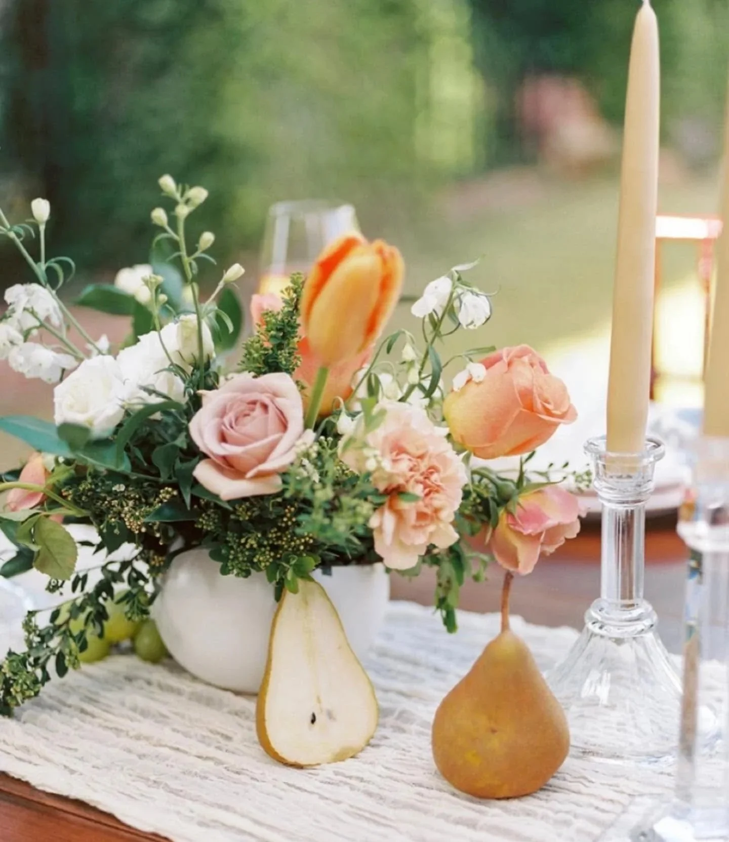 A floral table centerpiece with pink and peach roses, white flowers, and greenery in a white ceramic vase, next to a sliced pear, a tall yellow candle in a glass holder, and an unlit white candle in a tall glass holder. The background is blurred with greenery visible outside.