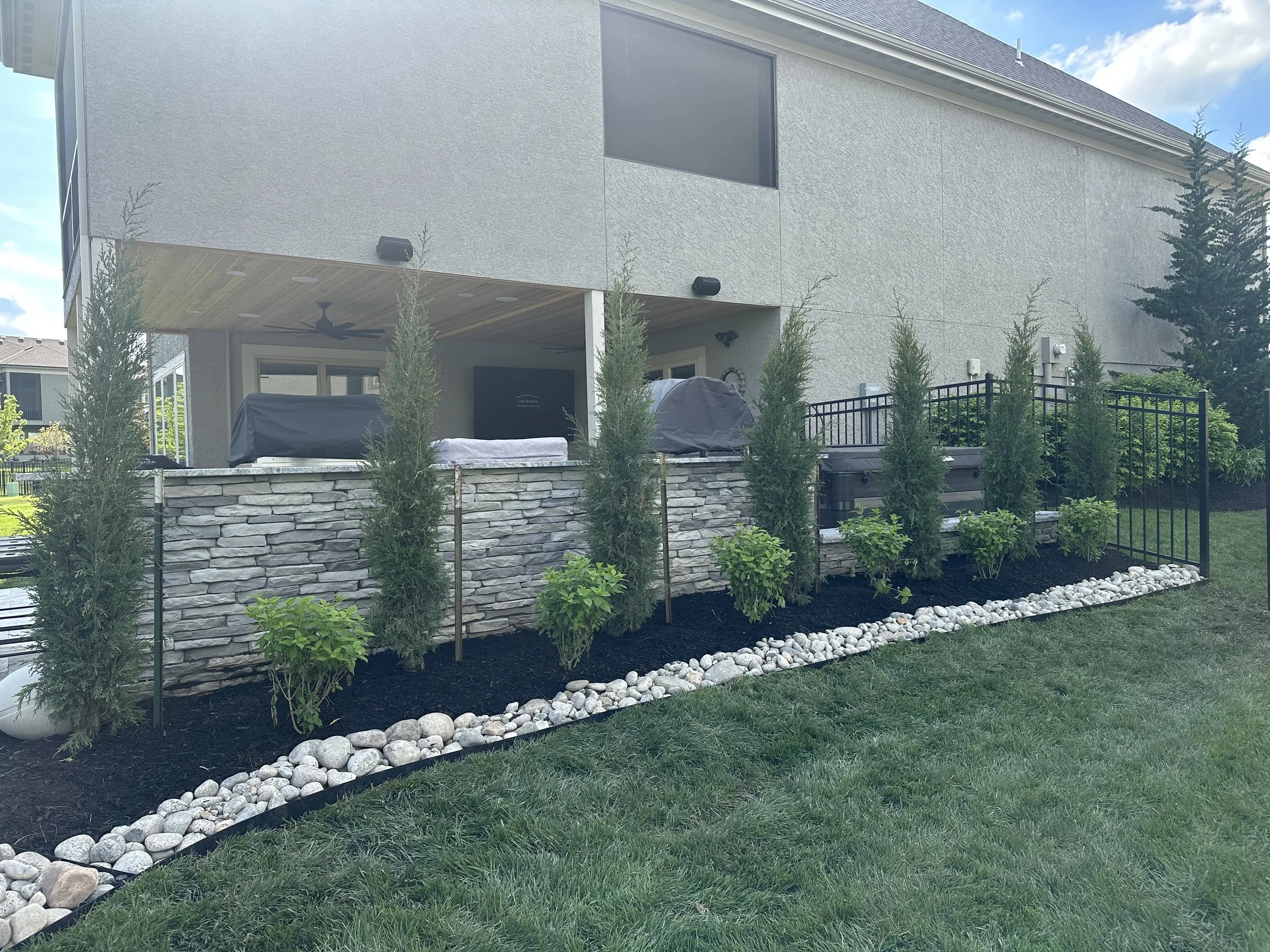 Backyard patio with stone wall, tall evergreen trees, and a fenced area. The house has a stucco exterior with a large window and a covered patio with a ceiling fan and black wall lights. Outdoor furniture and a grill are visible.