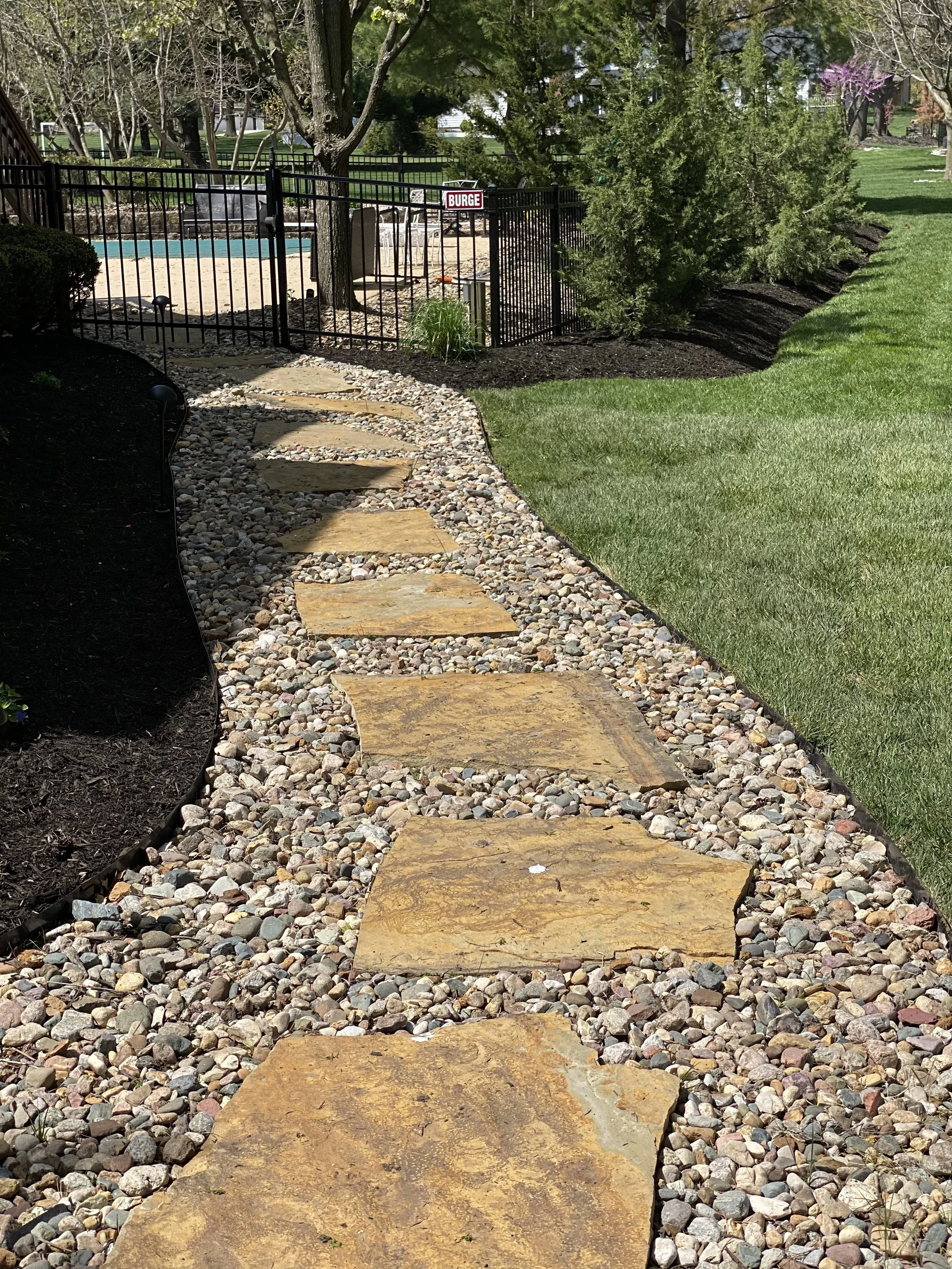A garden path made of large, flat stones goes through a gravel area with small, multicolored pebbles. There is a black metal fence surrounding a small backyard area with a pool, patio furniture, and trees with budding leaves and some small blossoms.