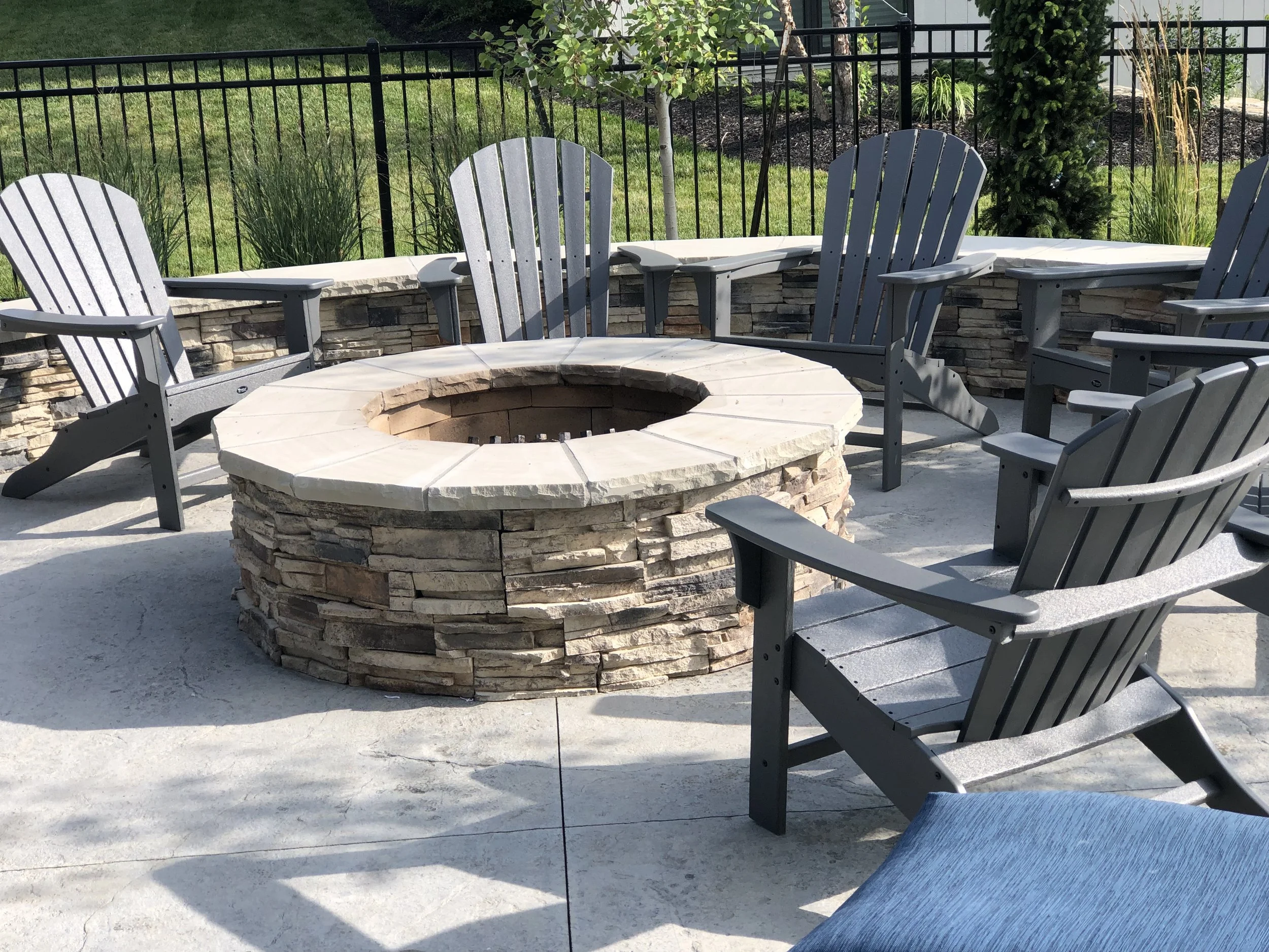 Outdoor patio area with a circular stone fire pit surrounded by multiple gray Adirondack chairs, lush green grass, plants, and small trees in the background, enclosed by a black metal fence.