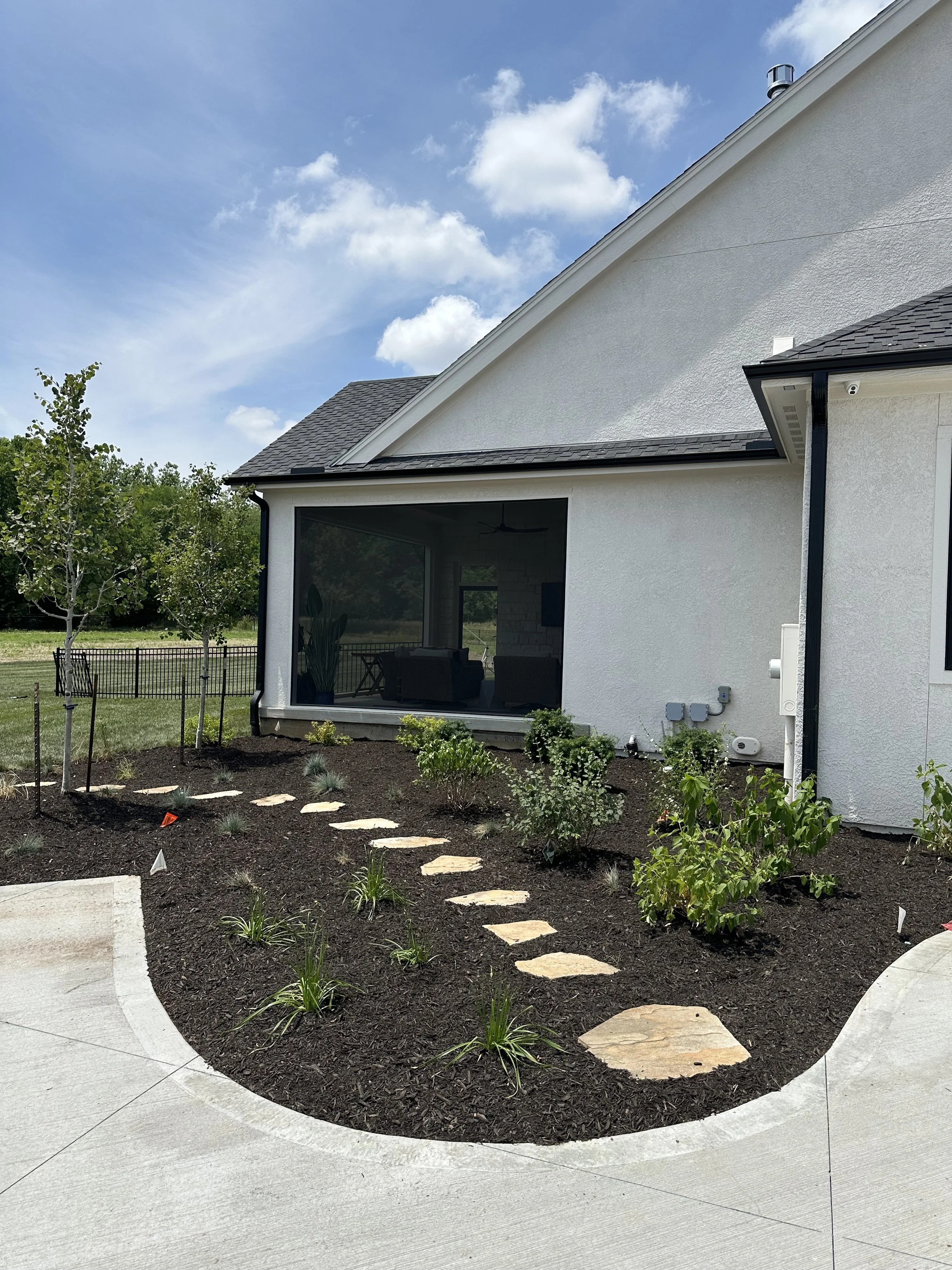 A backyard garden with newly planted small bushes and plants, a pathway of stepping stones, and a white house with a shaded porch, black gutters, and a screened area. End of the garden is a lush green field with trees and a blue sky with some clouds.